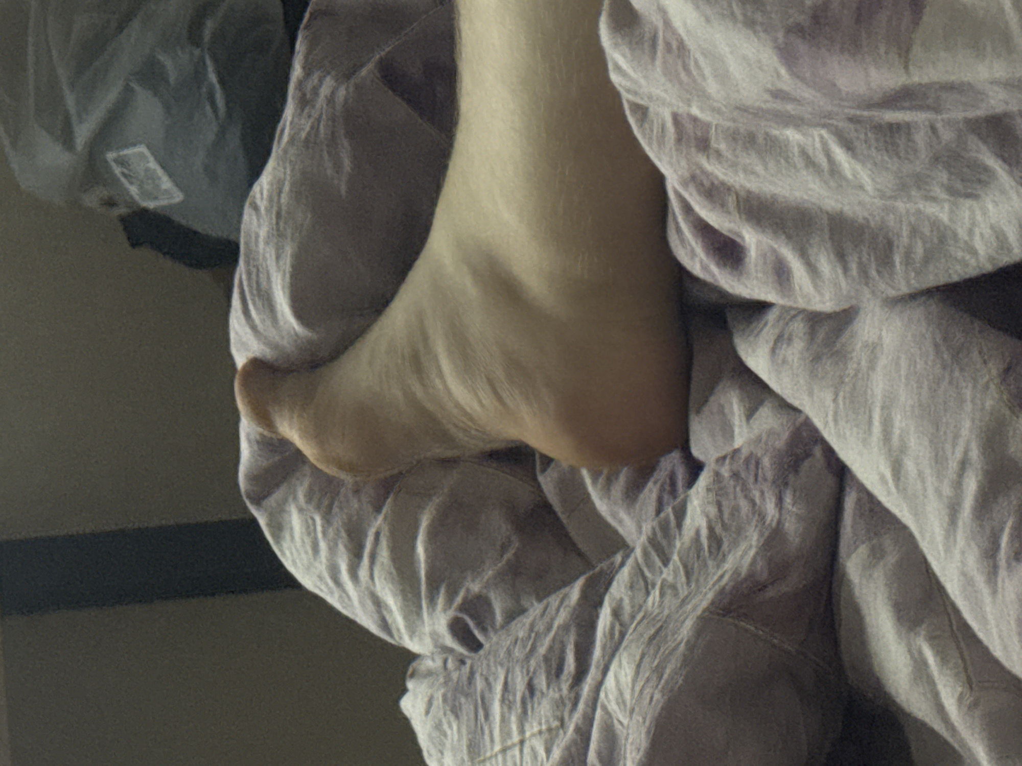 The image shows a close-up of a person's bare foot resting on wrinkled, light purple bedding. The foot appears to be positioned on its side, with the sole facing upwards. The surrounding bedding is textured and appears to be made of a soft fabric, possibly cotton or linen. In the background, out of focus, is a dark vertical object, and to the right, a blurry, large, light gray bag with a white label. The lighting is dim, suggesting an indoor setting, possibly a bedroom.
The image does not provide any information that would allow for the determination of its location in the city of Saratov, Russia.