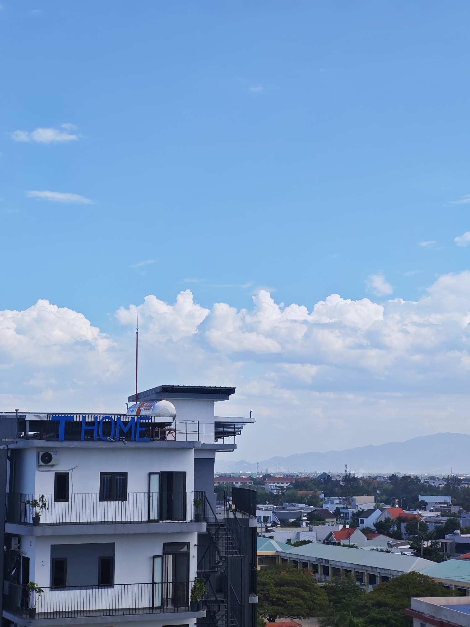This is a photograph taken from a high vantage point, likely a building, overlooking a city scene in Da Nang, Vietnam. The upper portion of the image is dominated by a bright blue sky with scattered white, puffy clouds.
In the mid-ground, a white multi-story building is prominent. The top of this building features blue lettering that spells out "T HOME". The building has several balconies with black railings and dark-framed windows. An outdoor staircase made of dark metal is visible on the right side of this building. An air conditioning unit is mounted on the exterior wall of the building. On the rooftop of the "T HOME" building, there appears to be a large, white cylindrical object, possibly a water tank, with some red markings. A slender, tall antenna or rod extends upwards from this rooftop area.
Below and beyond the "T HOME" building, a sprawling urban landscape unfolds. Various buildings with different colored roofs (some red, some green, some white) are visible, interspersed with lush green trees. In the distance, hazy blue mountains form the backdrop of the city. The overall impression is a sunny day in an urban environment.