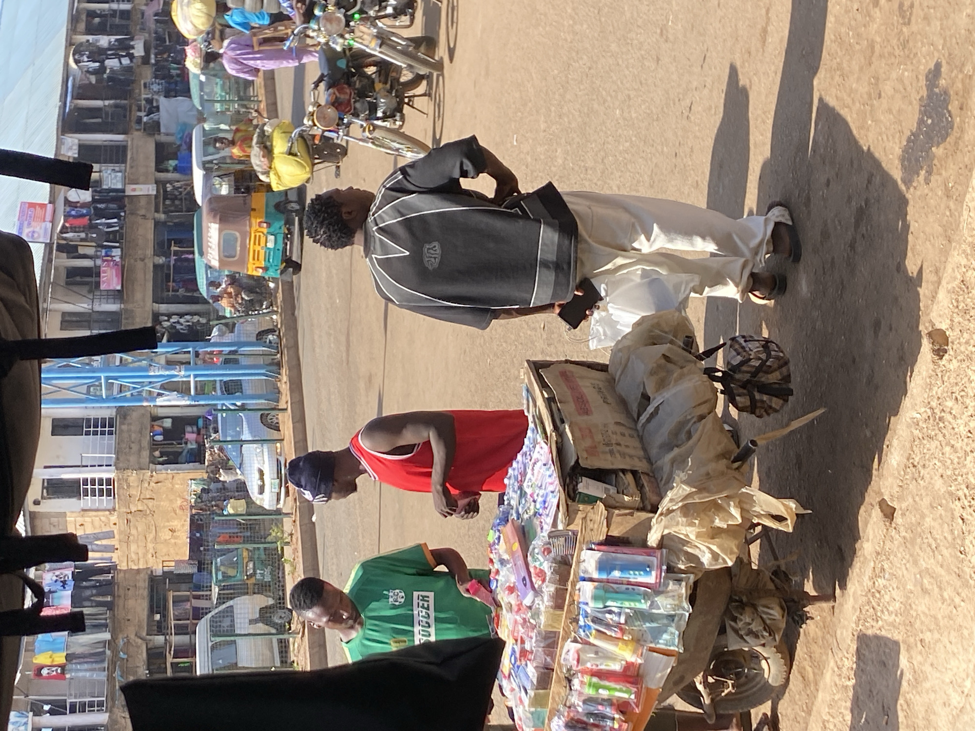 The image depicts a street scene in Bukuru, Nigeria. The photograph is oriented vertically. The environment is sunny, and the street is paved.
Two men stand near a cart laden with merchandise. One man wears a green soccer jersey, and the other wears a red tank top and a dark head covering. The cart displays various items, including pens, stationery, and other small goods.
A third man, wearing a black and white jacket and light-colored trousers, stands nearby. In the background, a row of buildings with varying architectural styles is visible. Some of the buildings have exposed brick and multiple windows. A few vehicles, including a tricycle taxi, are parked along the street. Several motorcycles are in the background.