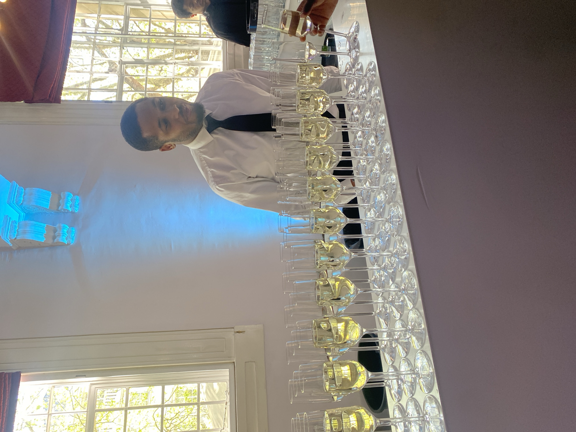 The image, taken in London, United Kingdom, depicts an indoor event setting, likely a reception or social gathering, focusing on drink preparation. A man, dressed in a white collared shirt and black tie, stands in the foreground behind a long white table. He looks directly towards the camera with a neutral expression.
The table is systematically arranged with numerous clear champagne flutes. Many of these glasses are filled with a pale yellow liquid, presumably champagne or sparkling wine, while others appear empty or partially filled, indicating an ongoing service. Further down the table to the right, another individual, mostly obscured and wearing a dark uniform, is seen actively pouring the beverage into a glass from a bottle.
The background features large, multi-paned windows with white frames, through which bright natural daylight illuminates the scene. Green foliage and tree branches are visible outside, suggesting an outdoor view during daytime. A red curtain is partially visible on the far left. On the left wall, a decorative white architectural element, possibly part of a mantelpiece or molding, is highlighted with a distinct blue artificial light, adding to the ambiance. The setting appears to be a refined venue, perhaps a function room, preparing for or in the midst of an event. There is no visible text in the image.