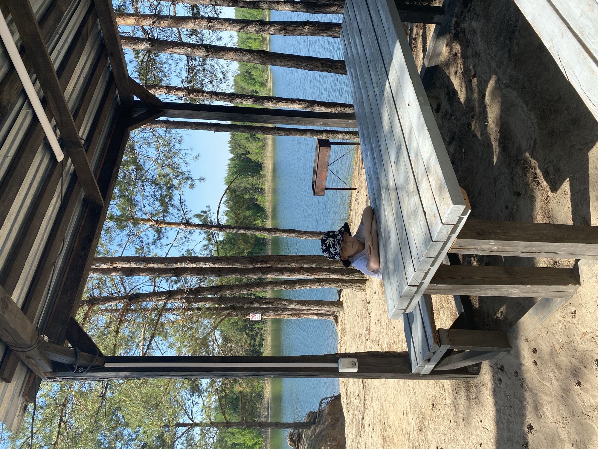 A medium shot shows a gazebo on a sandy shore with a lake in the background. A child wearing a hat is lying on a gray picnic table inside the gazebo. The gazebo is made of dark wood. Pine trees are visible along the shoreline, and there is a metal grill in the distance. A no-smoking sign is attached to one of the trees. An electrical outlet is visible on the post next to the sand shoreline. The image was taken in Novyi, Russia.