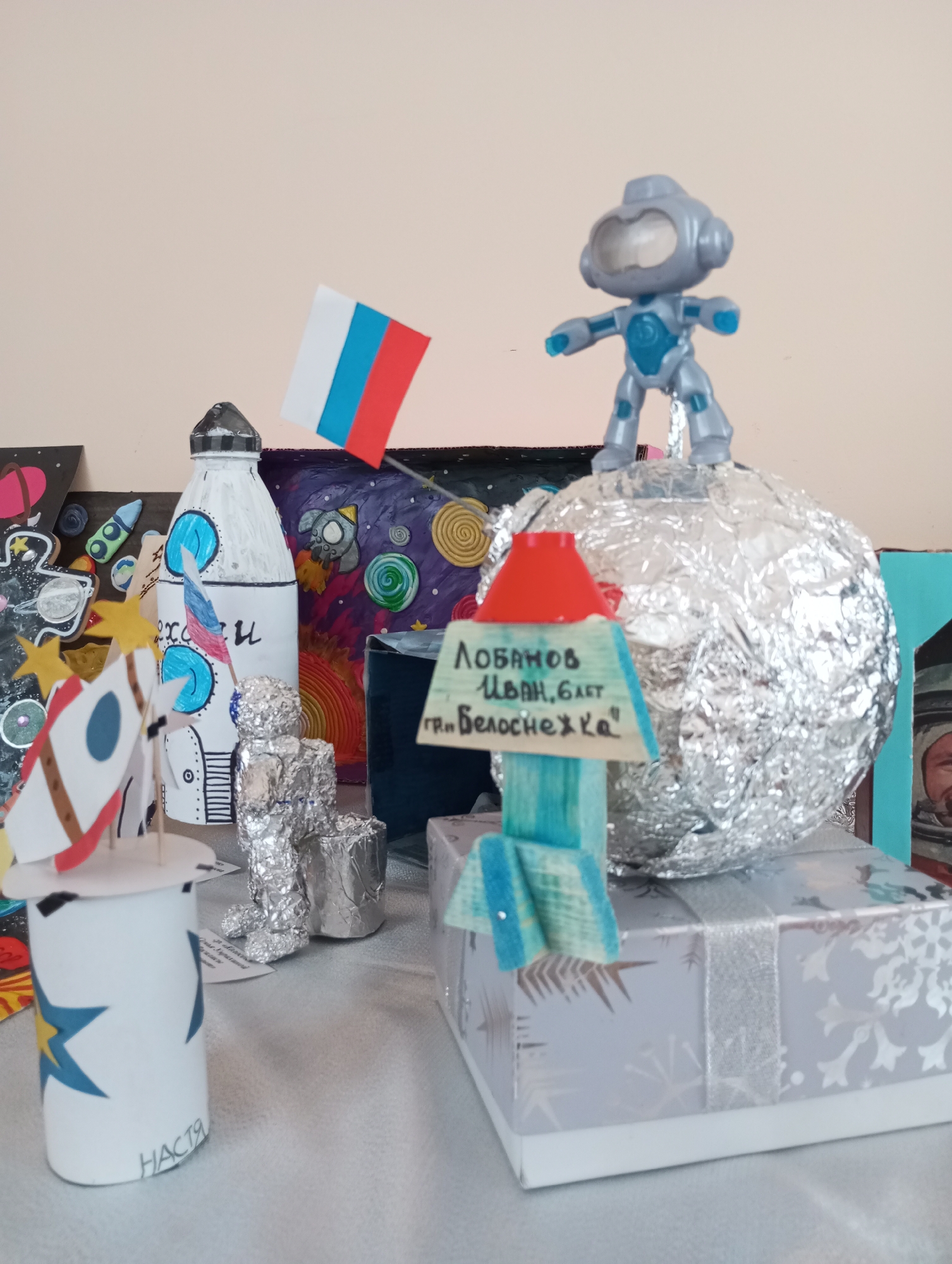 A tabletop display in an unknown location, possibly a city called "Unknown," features several handmade space-themed decorations. A rocket made from a plastic bottle and a paper rocket are visible. There is a figure covered in aluminum foil, presumably representing an astronaut, and a Russian flag. Another figure of an astronaut stands on a foil-covered sphere, representing a planet or moon. A sign with writing in Russian is partially visible. Other decorations and drawings are in the background. The overall impression is of a children's craft project or school display related to space exploration.