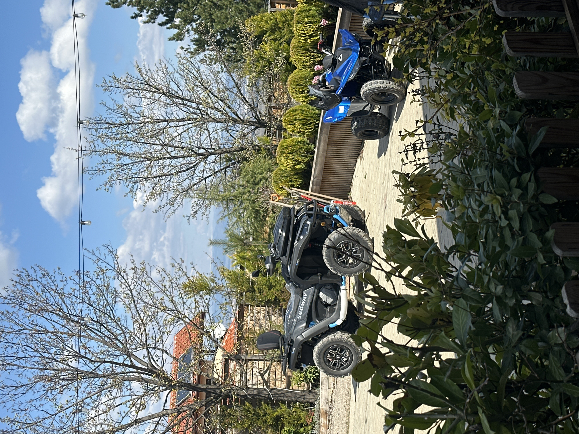 The image shows a sunny outdoor scene in Unknown, Unknown. The ground is paved with light-colored stones. In the foreground and center, there are two all-terrain vehicles (ATVs). One ATV is black and has the word "SEGWAY" visible on its side. It is positioned with its front facing the lower part of the image. Behind it, partially obscured, is a smaller, blue ATV. Various trees and bushes are scattered throughout the scene, creating a natural backdrop. A building with a red-tiled roof is partially visible through the trees on the left side of the image. The sky is blue with scattered white clouds. Power lines with lights are seen in the left of the sky.