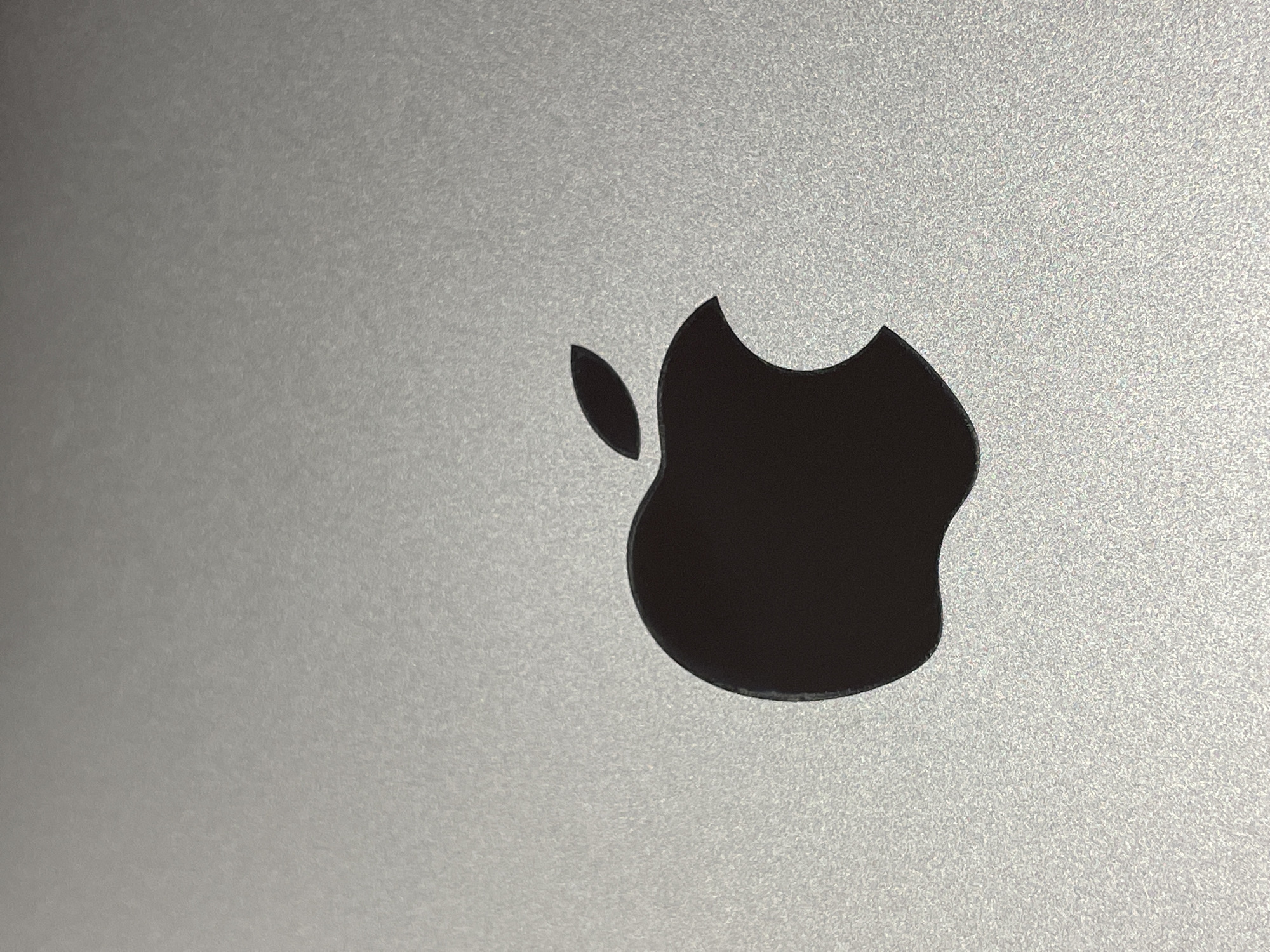 A close-up, high-angle shot features the iconic, dark gray or black Apple logo, complete with its distinct bite mark and a separate leaf element, prominently centered against a light gray, textured metallic surface. The surface, likely anodized aluminum, exhibits a fine-grained, slightly reflective texture, suggestive of a MacBook or iPad casing. Light reflects off the metallic finish, revealing subtle variations in brightness and a speckled appearance across the background, with the upper left area appearing slightly darker and the lower right subtly brighter. The logo itself has a smooth, matte finish, contrasting with the granular texture of its surroundings. No activity is depicted, and no text is visible. The scene is static, focusing purely on the product's design and material. The image provides no specific time of day or weather cues, indicating an indoor setting. This item is located in Kazan, Russia.