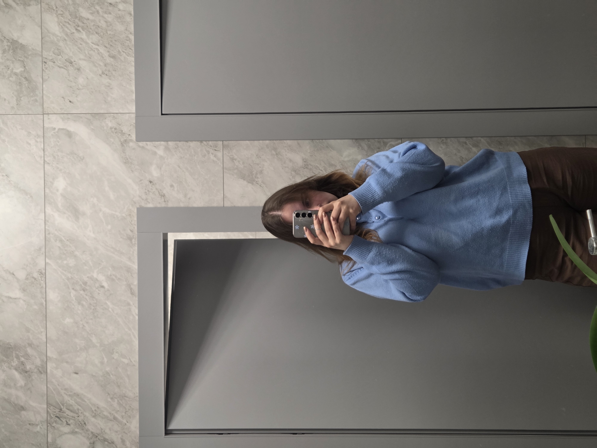 A person takes a photo of themselves in a mirror. The person is wearing a light blue sweater and brown pants. The background includes gray marble tiles and a gray cabinet. The location is Unknown, Unknown.