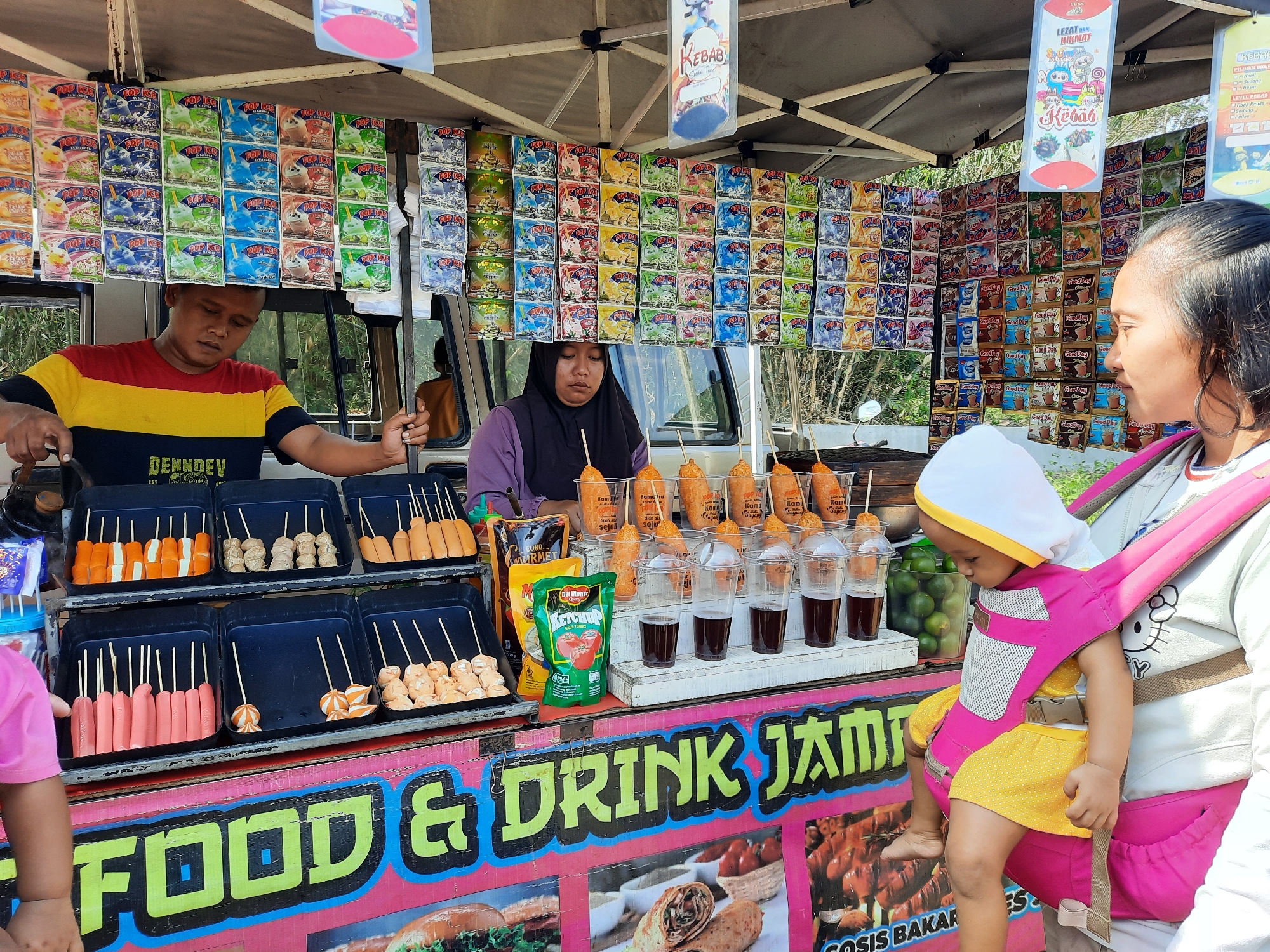 A street food vendor stall in Lugu, Indonesia, is depicted from a slightly low, eye-level perspective. The stall is crowded with various food items and beverages for sale, and two vendors are actively working. In the foreground, a woman is carrying a baby in a pink and yellow baby carrier. The baby, facing away from the camera and towards the stall, is wearing a white and yellow hat and a yellow skirt. The woman's right arm is visible, resting on the baby carrier.
Behind the woman, a male vendor with dark hair is wearing a red, yellow, and blue striped t-shirt with "DENNDEV" printed on it. He is leaning forward, appearing to prepare food. To his right, a female vendor wearing a purple shirt and a black hijab is also attending to the stall. She is holding a skewer of food.
The stall displays an array of colorful packaged snacks and drinks. Rows of "Pop Ice" flavored drink packets are hung prominently above. Various fried food items on skewers, such as sausages and fish cakes, are arranged in black trays. Cups of dark liquid, possibly iced coffee or soda, and a packet of Del Monte Ketchup are also visible on the counter.
The background shows lush green foliage, suggesting an outdoor location. A motorcycle is partially visible to the left of the stall. The stall itself is adorned with large, colorful signage that reads "FOOD & DRINK" in bold, stylized lettering. Additional images of food items, including grilled sausages and wraps, are printed on the stall's front panel. The overall scene captures the vibrant atmosphere of a local Indonesian street food market.