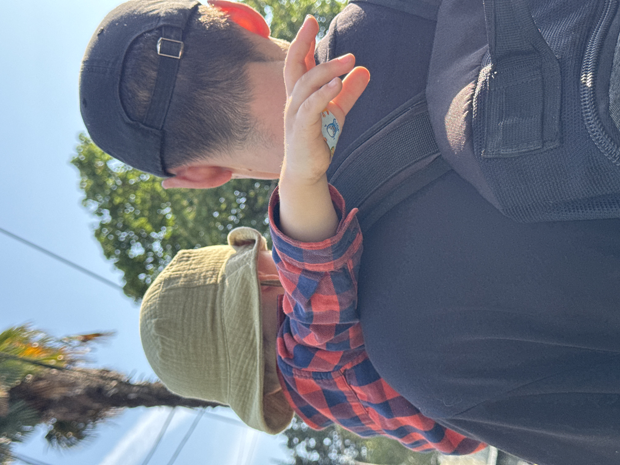 The image depicts a man carrying a child on his back. The man is wearing a black baseball cap and a black backpack. The child is wearing a green bucket hat and a red and blue plaid shirt. The child has a bandage on their hand. The background includes trees and a blue sky. Given the presence of palm trees, and judging from the color, it is likely daytime. It is not possible to determine whether the image was taken in Sochi, Russia from the information available.