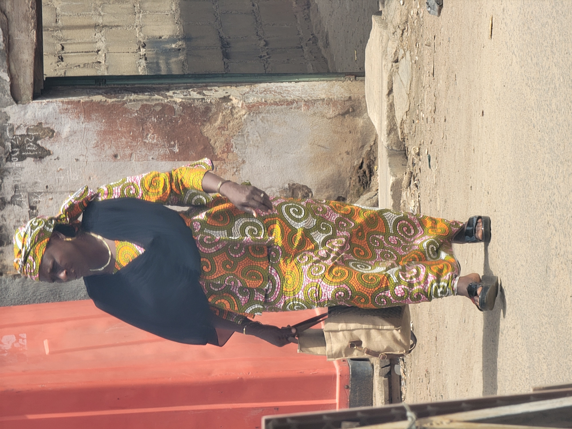 A woman stands outdoors in a sunny daytime setting in Maiburiji, Nigeria. She is dressed in a vibrant, long patterned garment, likely a dress, featuring swirling designs in shades of green, orange, pink, and white. This is paired with a plain black top underneath. A matching headwrap covers her hair, and she accessorizes with gold-colored hoop earrings and multiple gold bracelets on her wrists. She wears black platform sandals.
The woman holds a light-colored, possibly beige or khaki, shoulder bag or satchel with her left hand. She is positioned next to a tall, weathered wall with chipped paint revealing a rough, possibly brick or concrete, texture beneath. To her immediate left, a large, orange or red rectangular object, resembling a shipping container or metal structure, is prominently featured in the foreground. The ground she stands on is a light-colored, rough surface, likely concrete or packed earth, with small stones and debris scattered about.
Her gaze is directed forward and slightly downwards, with a neutral expression. The bright light and distinct shadows suggest it is a clear day. The clothing style and rustic background elements are consistent with the provided location context. No legible text is visible in the image.