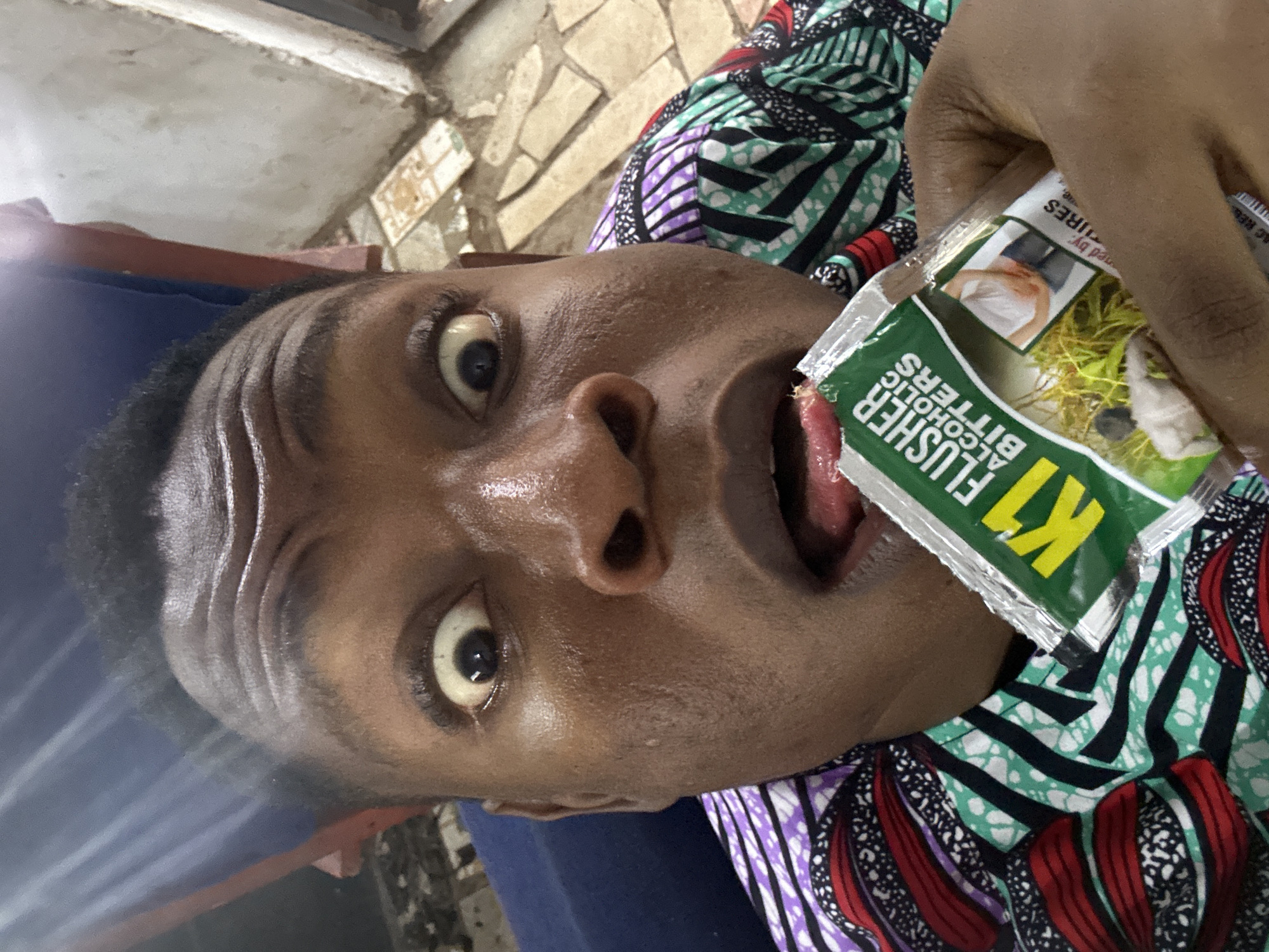 A close-up, slightly rotated image shows a person holding a sachet of "K1 Flusher Alcoholic Bitters" in front of their open mouth with their tongue protruding. The person has dark skin, wide eyes, and is wearing a colorful patterned shirt. The background is blurred and indistinct, revealing a hint of patterned wall behind. The location is Unknown, Unknown.