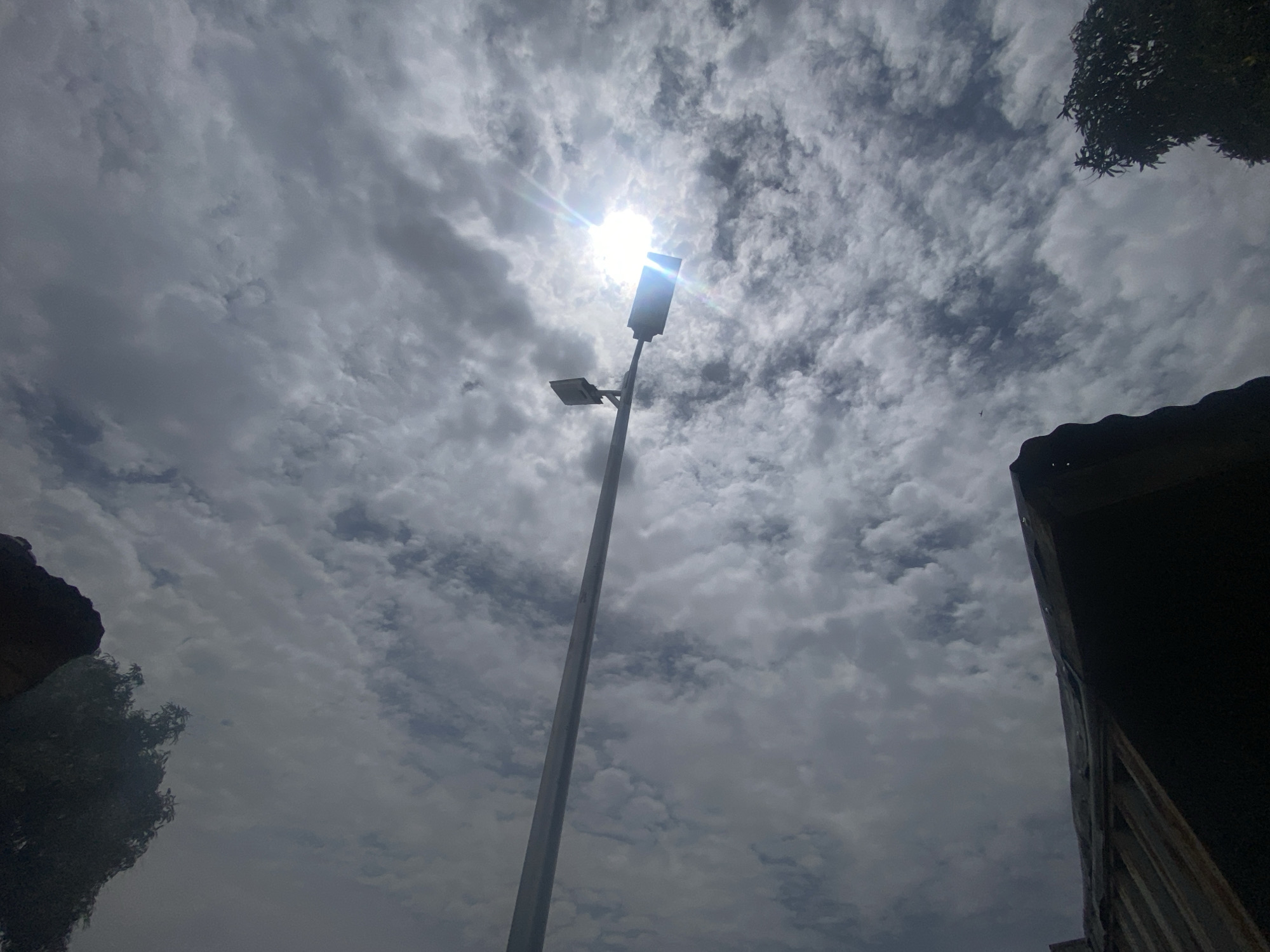 The image shows a view looking upwards towards a partly cloudy sky in Barden Yana, Nigeria. The sun is visible, partially obscured by a cloud, and shining through a street light. The street light is a tall, gray pole with two light fixtures. In the foreground, there is foliage and part of a building structure on either side of the image, partially obscuring the view. The sky appears overcast.
