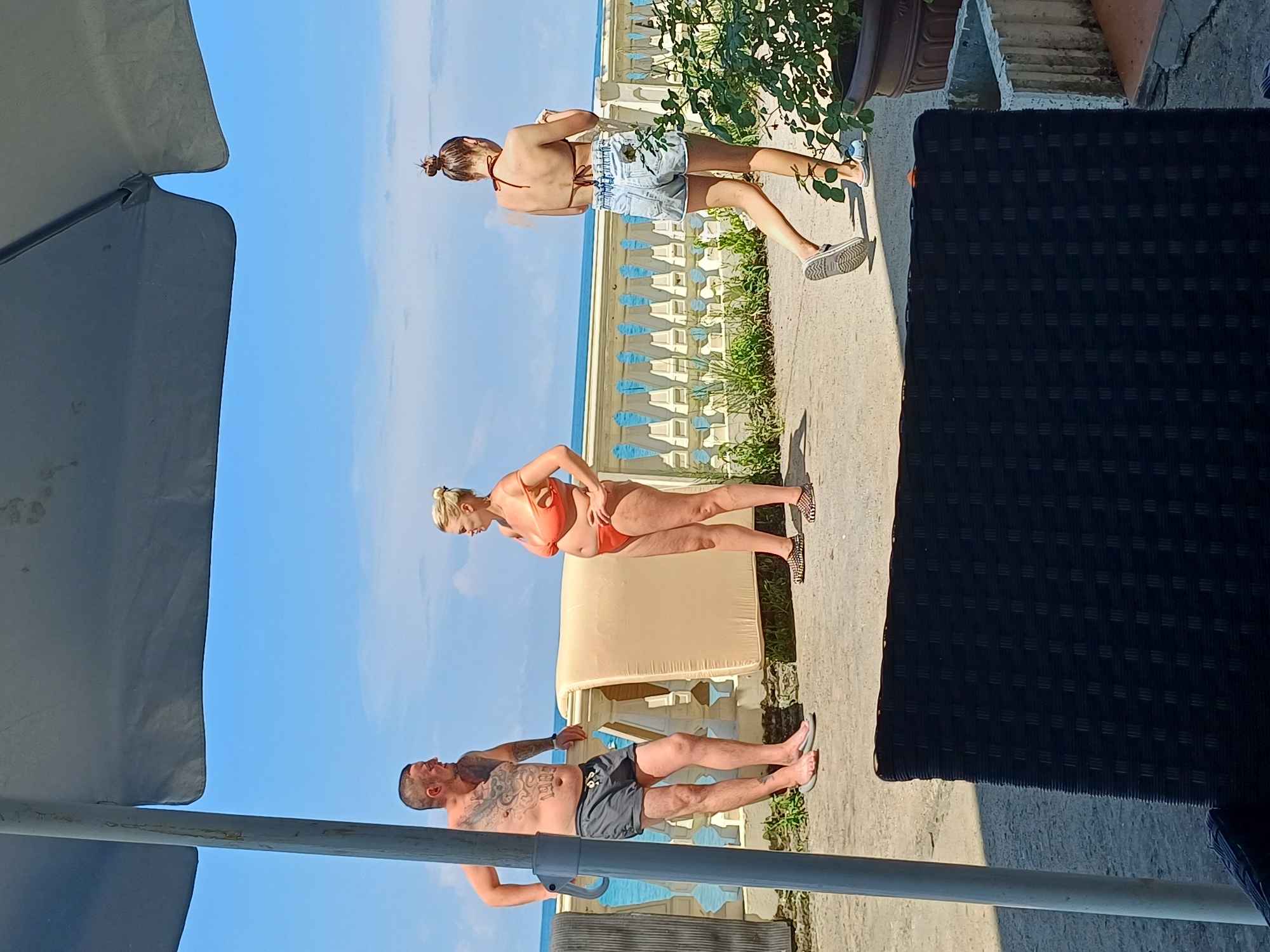 A man and two women are standing on a paved area near the Black Sea in Sochi, Russia. The man, on the left, wears grey shorts and no shirt, revealing tattoos on his torso. He leans against a white railing. The woman in the middle wears an orange bikini and is facing the man. The woman on the right, with her back to the camera, wears a brown bikini top and denim shorts, and appears to be walking away while holding something. Behind them, a white railing lines the edge of the paved area, and beyond that, the blue sea is visible under a clear sky. A large umbrella is partially visible in the foreground, casting a shadow. There is also a large cushion-like object leaning against the railing. A potted plant with green leaves is on the right side of the frame.