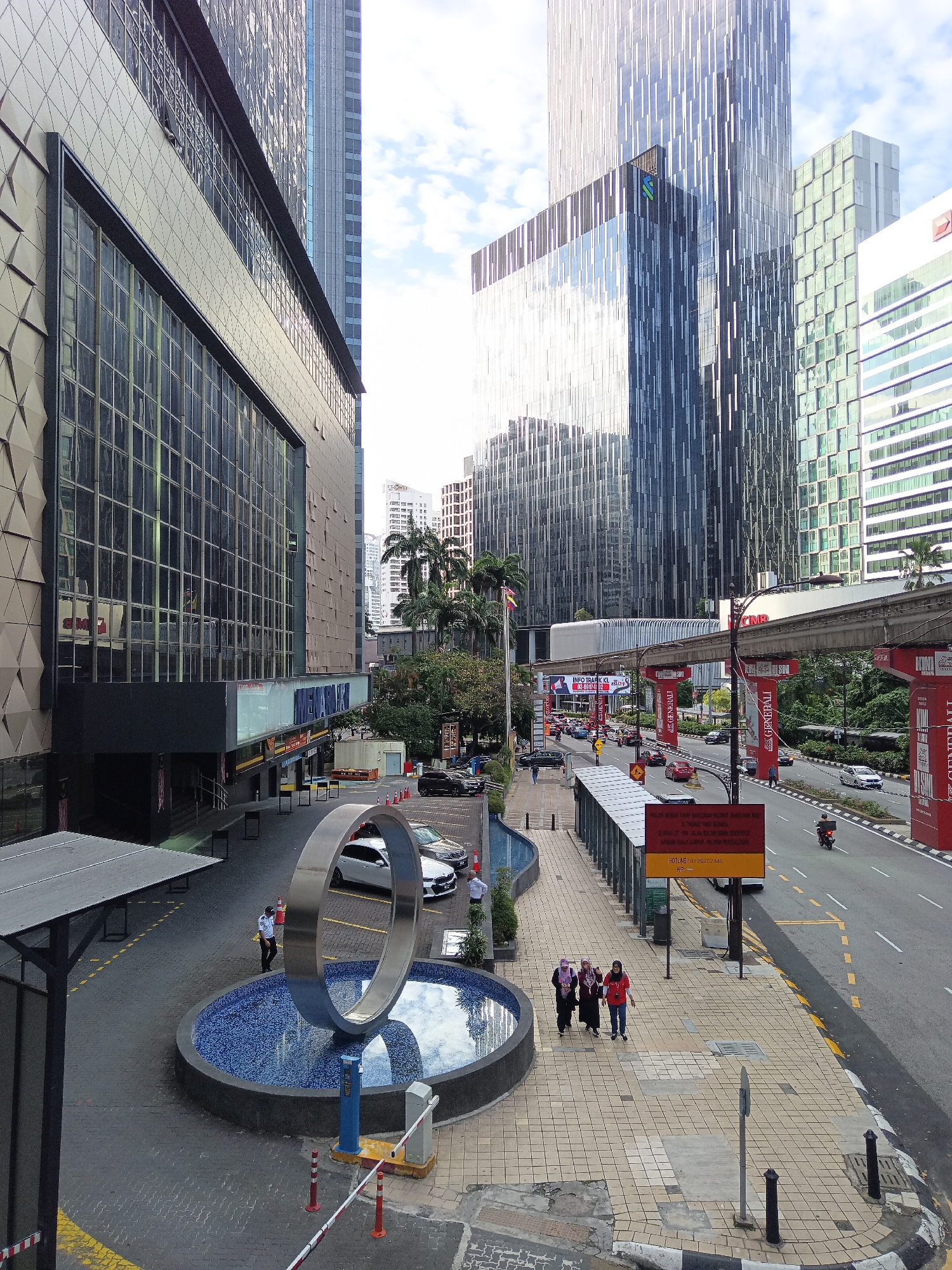 The image captures a street scene in Kuala Lumpur, Malaysia, likely in the vicinity of a commercial area or shopping mall. In the foreground, a large, modern sculpture resembling a silver ring sits within a circular fountain with a blue mosaic base. A security guard in a light blue uniform stands near the sculpture, and a few people are walking on the patterned sidewalk. Beyond the fountain, a road is visible with several cars and a motorcycle. Overhead, a monorail track runs across the scene, supported by red pillars adorned with banners. Tall, modern buildings with glass facades dominate the background, reflecting the sky and clouds. The architecture suggests a contemporary urban environment. Several traffic signs, including a red and yellow informational sign, are posted along the roadside. The overall impression is of a busy, well-developed urban landscape.