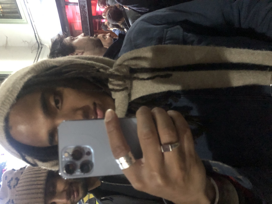 A selfie taken by a person wearing a beige hat, scarf, and blue jacket. The person has dreadlocks and is wearing two rings on their hand holding the phone. In the background, there are other people, some of whom are also looking at the camera. The background seems to be indoor. The lighting is slightly dim.