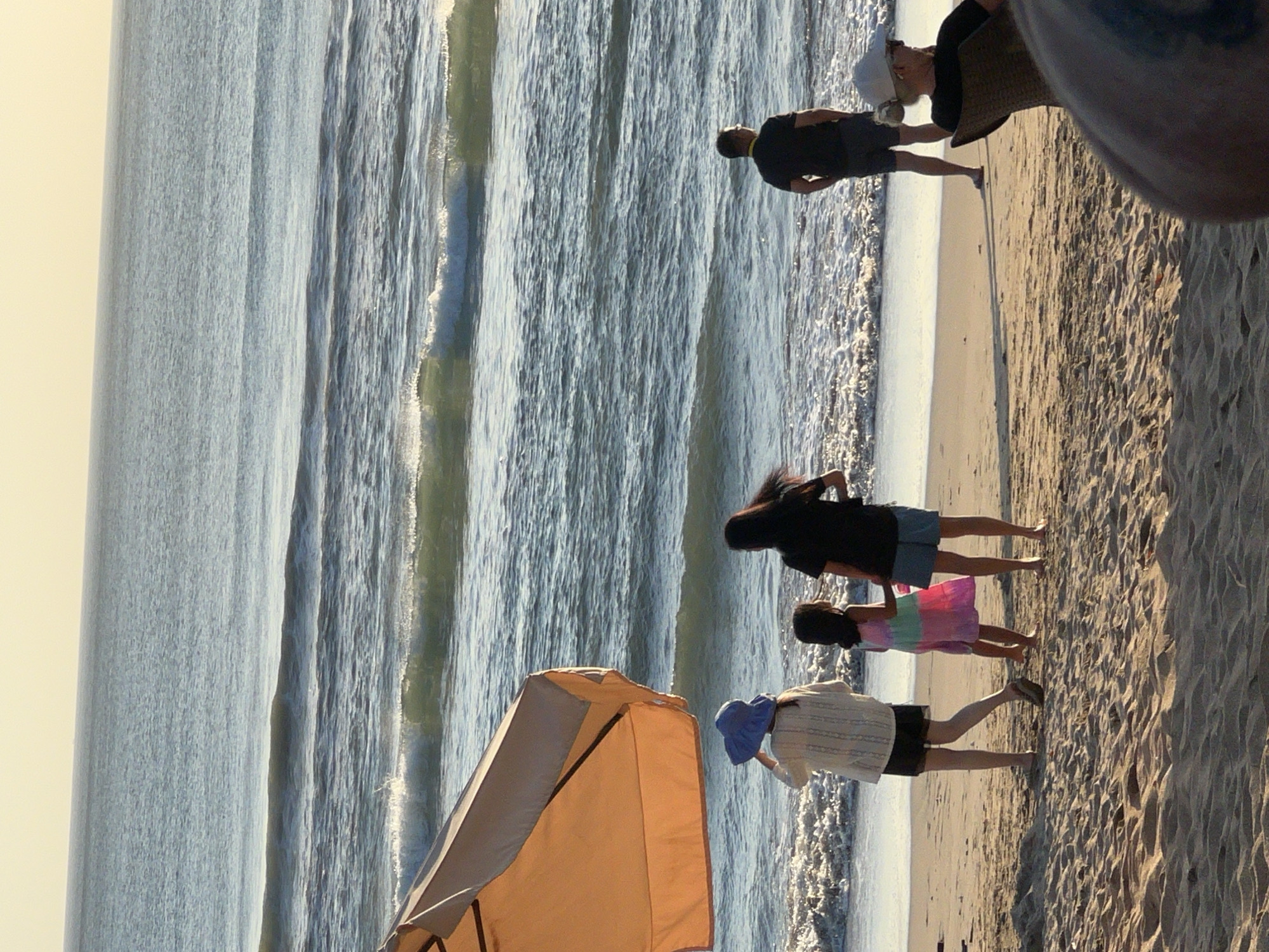 The image depicts a serene beach scene in Kuta, Indonesia, bathed in the warm glow of the setting sun. The vast expanse of the Indian Ocean stretches towards the horizon, with gentle waves lapping at the shore. In the foreground, a vibrant orange beach umbrella provides a splash of color against the golden sand.
Several individuals are scattered across the beach, enjoying the coastal ambiance. Two adults and a child stand near the water's edge, their backs to the camera, seemingly engaged in conversation or admiring the ocean view. Further down the beach, a man stands looking out at the sea, his silhouette sharp against the bright water. To the right, a woman is seated in a chair, facing away, her hat providing a cool shade.
The sand is marked with the footprints of those who have walked there, adding a sense of transient presence to the scene. The overall atmosphere is one of tranquility and relaxation, capturing the essence of a perfect day at the beach.