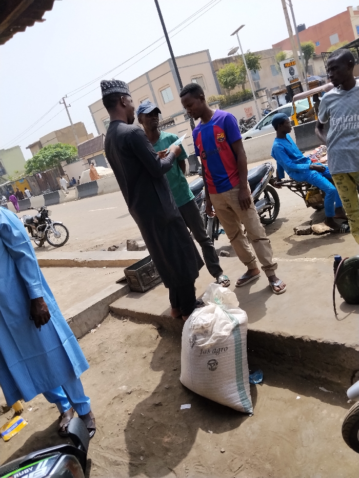 This image depicts an outdoor street scene in Fagge D2, Nigeria, during daylight hours. In the foreground, partially visible on the left, are the lower half of a person wearing a light blue traditional garment and dark sandals, next to part of a black and green motorcycle. On the ground nearby, a white sack labeled "fas agro" with a circular logo is present.
In the mid-ground, a group of three men stands on a raised concrete platform. The central figure wears a black traditional tunic and a black cap, holding a white rectangular object, possibly a phone or small book, and appears to be showing it to the two men beside him. To his right, a man in a green t-shirt and blue baseball cap observes the object. To his left, a man in a blue and red FC Barcelona jersey and tan pants also looks at the object. Behind this group, a silver scooter is parked.
To the right of the central group, another man in a grey t-shirt with "Emirates FLY BETTER" text and patterned pants stands, observing the group. Further right, a man in a bright blue traditional outfit sits on the cargo bed of a motorized tricycle (keke-napep), facing away from the primary interaction.
The background shows a paved street with light vehicle traffic, including cars and motorcycles. Several multi-story buildings are visible, some with satellite dishes or antennae. One building displays a partial sign with "NASTE." Utility poles and power lines traverse the scene, with a solar-powered streetlight also present. The ground is a mix of concrete, dirt, and dust. The scene is illuminated by strong overhead sunlight, casting distinct shadows.