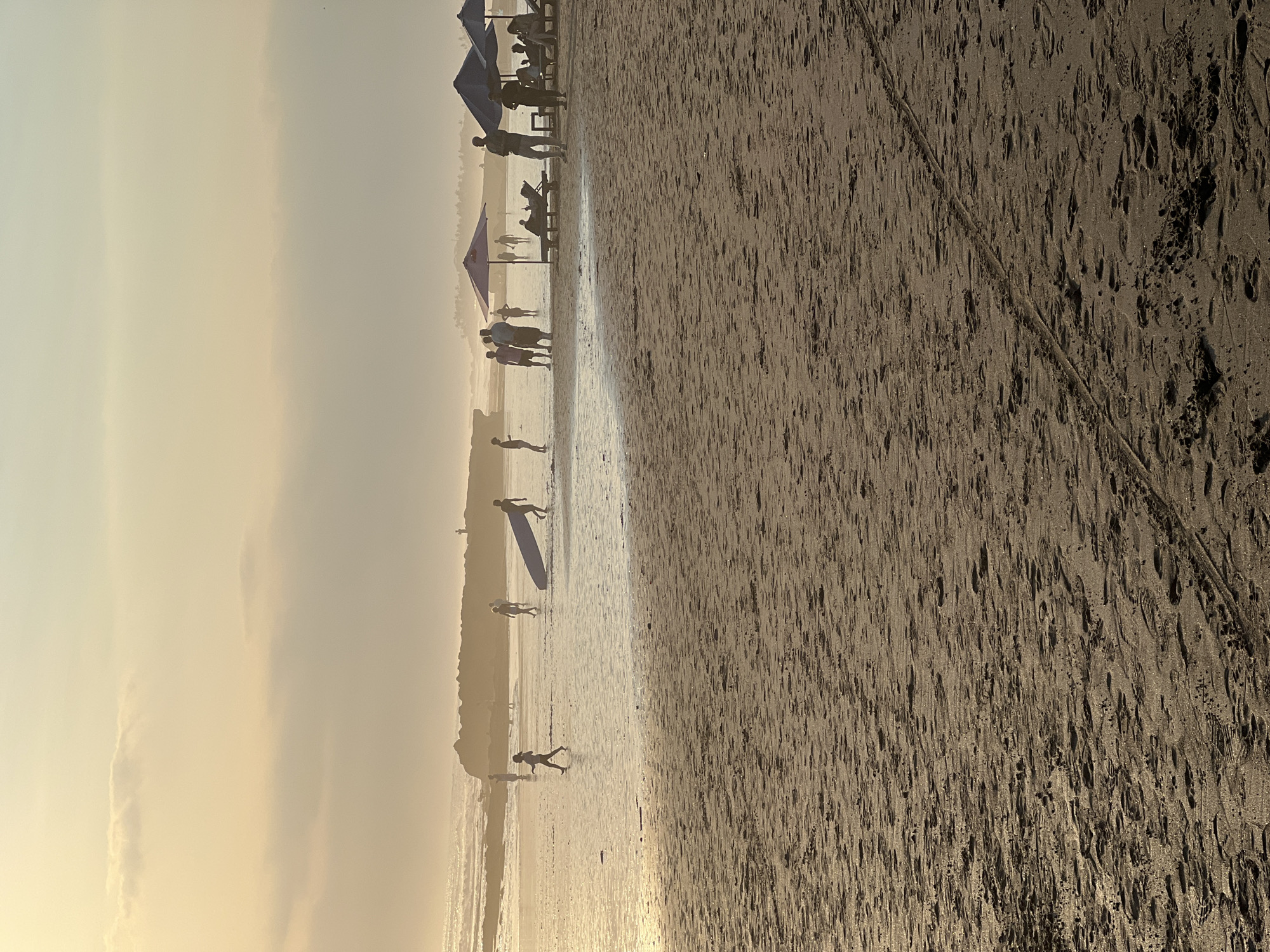 The image depicts a beach in North Kuta, Indonesia, during a sunset. Silhouettes of people are visible on the wet sand near the waterline, with some walking and one carrying a surfboard. In the background, a rock formation extends into the ocean, and a few individuals are standing on it. Further back, beach umbrellas and lounge chairs are set up on the drier sand, with more people relaxing. The sky is a soft gradient of yellow and orange, with subtle clouds, indicating the setting sun. The sand in the foreground is textured with numerous footprints.