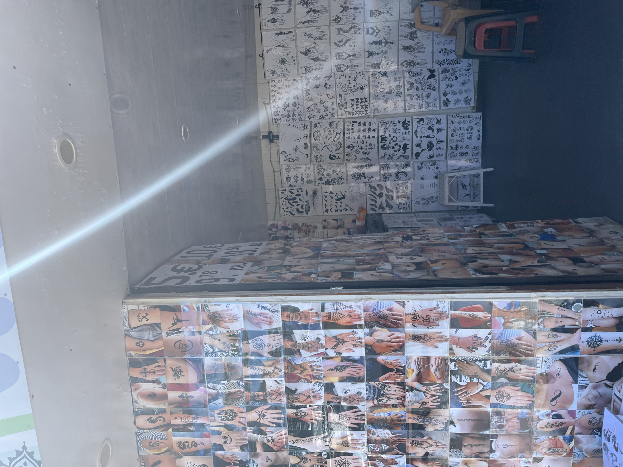 This media file shows a display of henna tattoo stencils and examples, likely at an outdoor market or stall in Nesebar, Bulgaria. The scene is a close-up view of the display, with a bright light source creating a strong glare across the left side of the image, suggesting it's daytime. The stencils, in black and white, are arranged in grids on sheets of paper, showcasing various designs such as flowers, butterflies, tribal patterns, and animals. Below the stencils, a collection of photographs displays finished henna tattoos on people's hands, arms, and other body parts. These photos offer a visual guide to the intricate and diverse artistic possibilities of henna.
The overall impression is of a vendor showcasing their artistry and offerings to potential customers. The vibrant display of designs and applied tattoos implies a busy or active setting where people might be looking for temporary body art. The backdrop appears to be a wall, possibly part of a stall or a building. The presence of a red chair or stool and some wooden elements further suggests a commercial or exhibition space. The overall mood is one of artistic presentation and visual appeal, designed to attract attention and encourage custom. No specific time of day or weather conditions are strongly indicated beyond the presence of bright light. The location context of Nesebar, Bulgaria, is provided externally, and the visual elements are consistent with a tourist-oriented market or shop.
