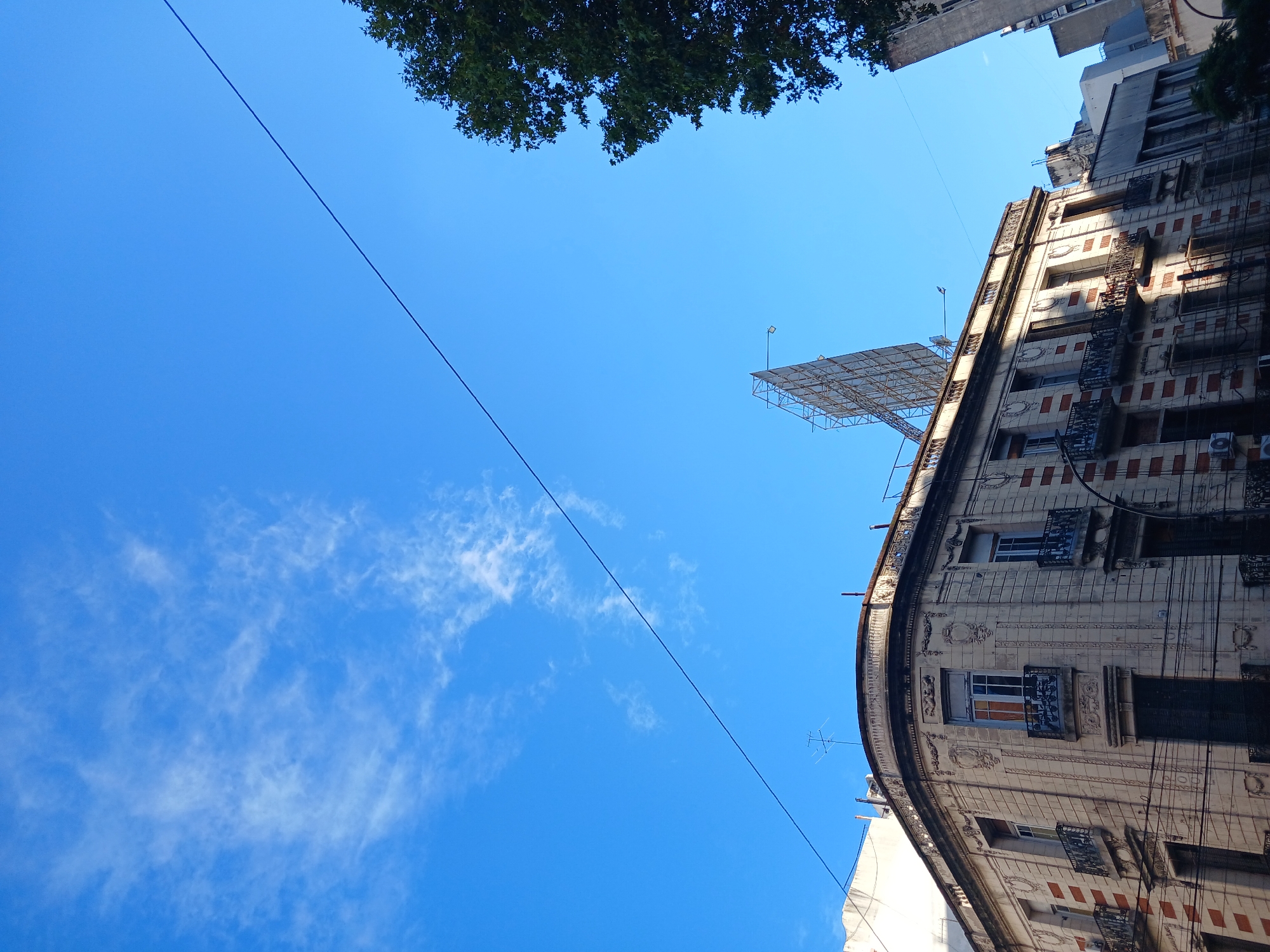 An upward-looking view captures a vibrant blue sky above buildings in Buenos Aires, Argentina. The main subject is a multi-story building, likely an apartment or office block, with an ornate and slightly curved facade that dominates the right side of the frame. Its architecture features intricate decorative molding, numerous windows, some with metal railings, and visible air conditioning units on its exterior. Strong sunlight casts distinct shadows on parts of the building, highlighting its textured surface and intricate details.
Above the main building, a large metal framework, possibly a billboard structure or a rooftop installation, is visible against the clear sky, with a single light fixture extending from it. To the far left and top of the image, the dark green canopy of a tree adds a natural element to the urban scene. Another, plainer building can be seen peeking in from the top left corner.
A thin black power line or cable stretches diagonally across the bright blue sky, from the bottom left towards the upper right, bisecting the frame. The sky is mostly clear, but delicate wisps of white clouds are scattered across the lower left portion. The scene depicts a bright, sunny day, typical of an urban environment. No people or visible text are present in the image.