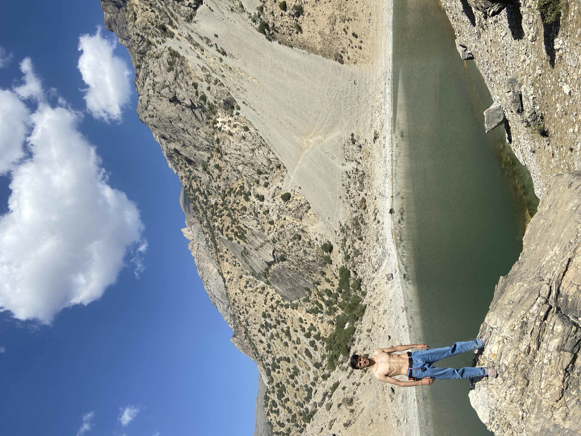 A shirtless young man in blue jeans and sandals stands on a rocky outcrop overlooking a serene, dark green lake. He is positioned in the lower left quadrant of the frame, facing the camera. Behind him, the landscape dramatically rises with a steep, rocky mountain face dotted with sparse vegetation. A vast, clear blue sky with scattered white clouds stretches across the upper portion of the image. The scene depicts a natural, mountainous environment, characteristic of the Ghazza region in Tajikistan.
