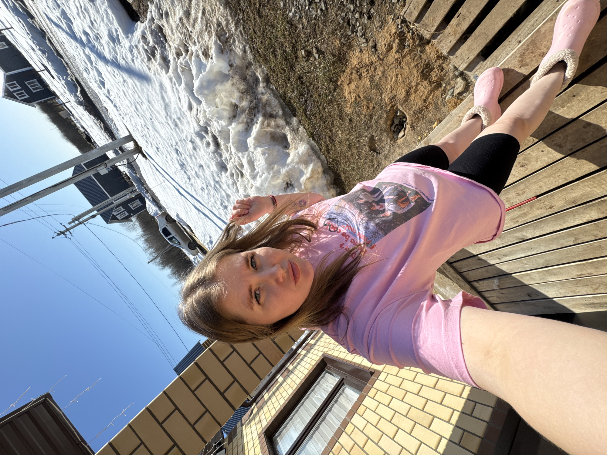 An outdoor selfie captured in Gornyi (Dvurechenskoe S/P), Russia, depicts a fair-skinned female subject with long brown hair, wearing a pink t-shirt with a graphic print, black shorts, and pink fur-lined clogs. Her left hand is raised, touching her hair. A small tattoo is visible on her left forearm. She gazes directly at the camera.
The subject is positioned on a wooden slatted deck. To her immediate left is a building with light yellow brickwork and a dark-framed window, partially covered by a beige awning. The background reveals a late winter or early spring scene with patchy snow cover melting into visible brown earth and sparse dry grass. Utility poles with overhead wires traverse the scene. Two dark-colored, two-story houses with white window trim are present in the mid-ground, beyond the snow. A light-colored sedan is parked near one of these houses. Bare trees are visible in the distant background against a clear blue sky. Bright sunlight illuminates the scene, creating distinct shadows.