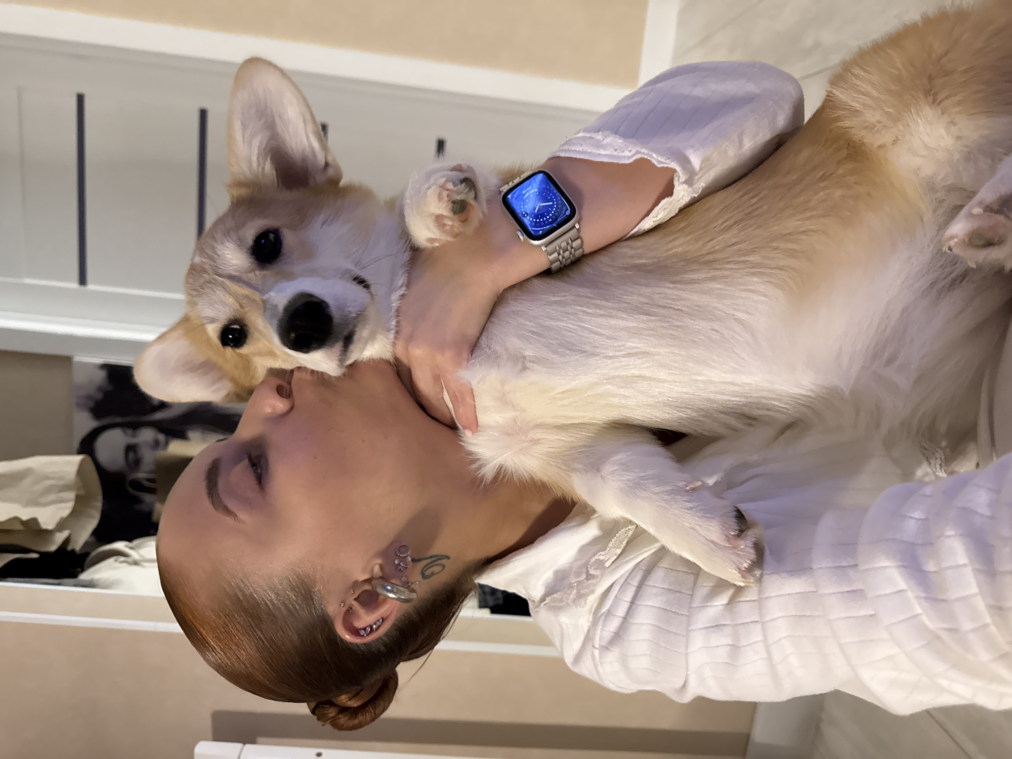 An indoor vertical shot captures a woman affectionately embracing a Corgi dog. The woman, with her hair styled in a sleek bun, is seen with her head turned, seemingly kissing or nuzzling the dog's head/neck area. She wears a white long-sleeved top with a ribbed or smocked texture. On her left wrist, she sports a smartwatch with a silver link bracelet. The watch face is illuminated in blue, displaying the digital time "10:01", the date "WED 10", and various small complications including temperature (22 degrees C), heart rate (92 bpm), and activity rings.
The Corgi, with its distinctive light brown and white fur, looks directly towards the camera, its ears perked up. Its front paws are visible, one resting near the woman's arm. The woman has multiple ear piercings, including studs, hoops, a heart-shaped piercing, and a larger circular one, along with a swirling tattoo visible on her neck/jawline area.
The background is an indoor setting, likely a home, with parts of a white wall or furniture visible. Behind the dog's head, there's a light-colored surface with two dark vertical lines. Behind the woman's head, a blurry, possibly framed, black and white portrait of a person can be discerned on the wall. The overall mood is one of warmth and affection between the owner and her pet.
The location context for this media file is Chaikovsky, Russia.