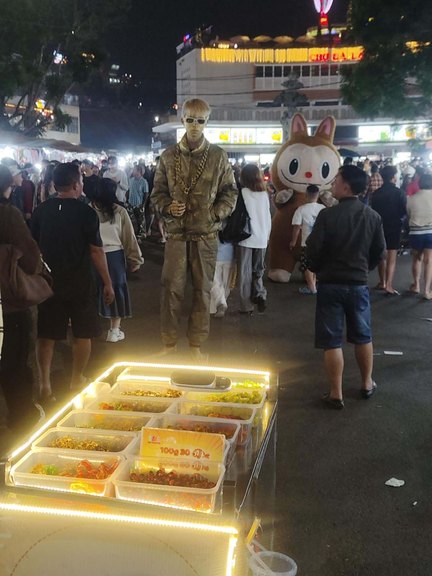 The image shows a night market scene in Da Lat, Vietnam. In the foreground, a vendor's stall displays various colorful candies and snacks in clear plastic containers, illuminated by bright yellow LED lights. Behind the stall, a mannequin dressed in camouflage clothing and a large gold chain stands prominently. To the right of the mannequin, a costumed character resembling a cartoon rabbit is visible. A crowd of people, appearing to be locals and tourists, are browsing the market. In the background, buildings with illuminated signs suggest a bustling urban environment. The overall impression is of a vibrant and lively night market.