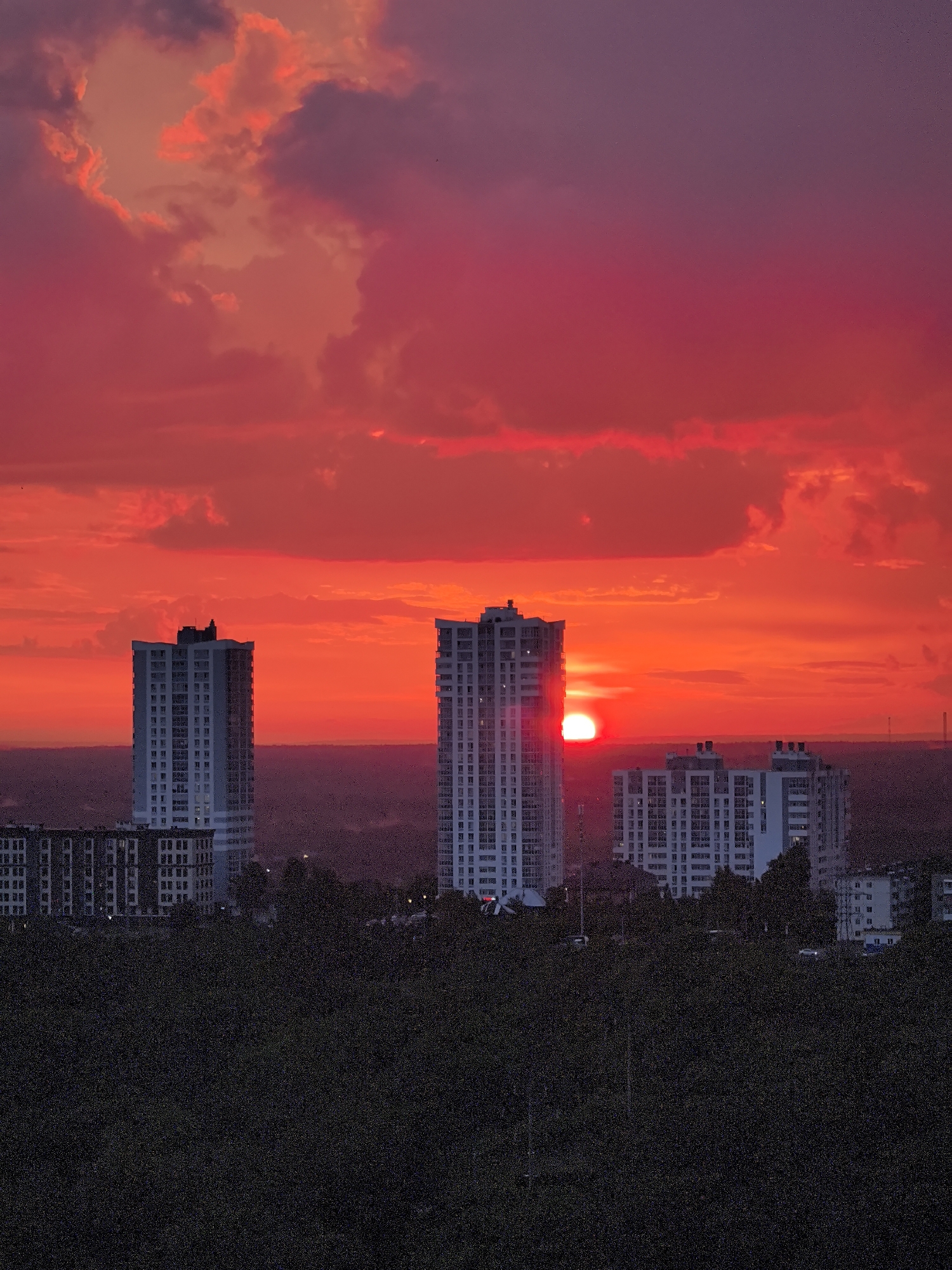 This is an aerial photograph of a city skyline at sunset, featuring tall residential buildings and a fiery red sky. The image depicts the Kamsky region of Perm, Russia. The foreground is dominated by a dark, dense line of trees, likely a park or forested area, that obscures the lower sections of the buildings.
Several high-rise buildings are visible. The most prominent are two tall, white apartment blocks with numerous windows. One building, on the left, is slightly taller and has a distinctive architectural feature at its top, possibly a ventilation system or decorative element. Another similar building is located to the right of the first, and slightly behind it. In the middle ground, there are several other, slightly shorter apartment buildings, also appearing to be residential complexes, with a more uniform façade. Further in the distance, a line of low-rise buildings is visible, and beyond that, the faint outline of more distant structures, suggesting a larger urban sprawl.
The sky is a dramatic and dominant feature of the image. It is filled with intense shades of red and orange, indicative of a sunset. Large, fluffy clouds are illuminated by the setting sun, creating a vibrant and almost surreal atmosphere. The sun itself is visible as a bright, glowing orb peeking out from behind the left-hand tall building, casting a luminous glow on its façade and the surrounding sky. The overall impression is one of a striking natural phenomenon occurring over an urban landscape. There are no people or vehicles clearly identifiable in the image.