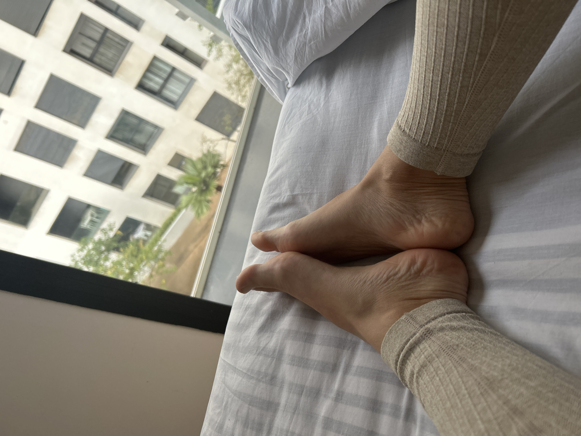 A close-up shot reveals a person's legs resting on a bed covered with a white and gray striped sheet. The person is wearing ribbed knit leggings in a neutral beige color. The soles of their feet are visible. A window behind the bed shows a tall building with many windows and a palm tree outside. While it's not possible to conclusively identify the location as Casablanca, Morocco from the image alone, the architectural style of the building and the presence of a palm tree are consistent with the region.