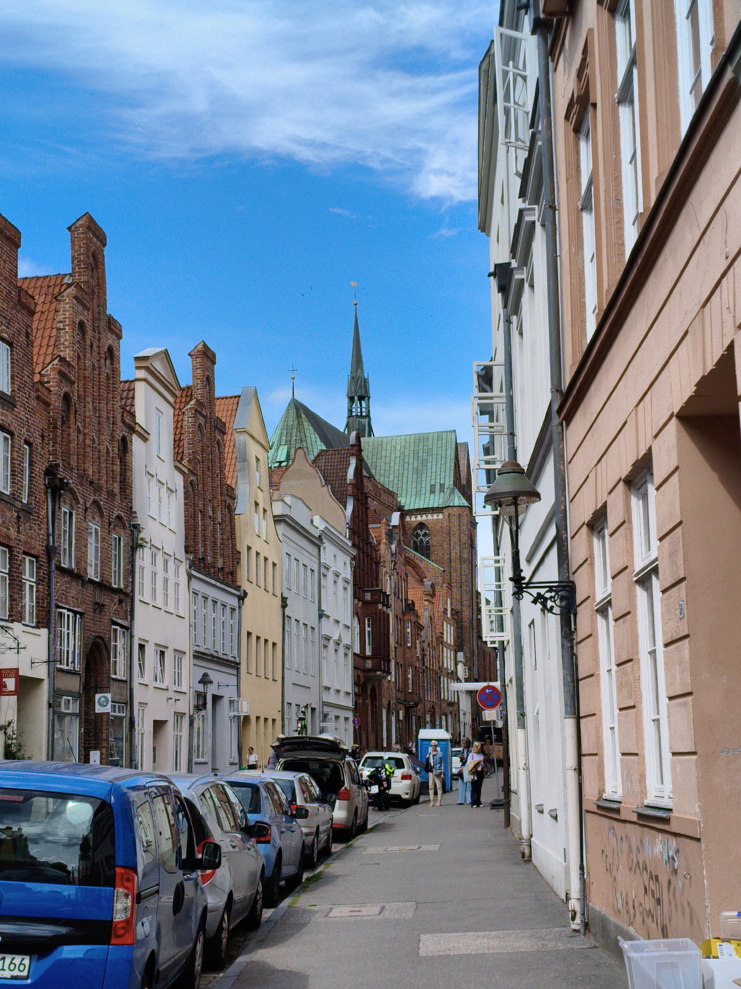 The image depicts a street scene in Lübeck, Germany, characterized by its historic architecture. On the left, a row of tall, narrow brick buildings with red tiled roofs and numerous windows line the street. These buildings have distinctive stepped gables. A church spire with a green roof is visible in the background. On the right, a light brown building with white-framed windows stands close to the sidewalk, with a lamppost attached to a pipe running down its facade.
The street is paved with concrete and lined with parked cars. The foreground features a bright blue van, followed by several other cars in various shades of blue, grey, and white. Some of these cars have their trunks open. A few people are visible on the sidewalk, including a woman in a white shirt and jeans, and another person standing near a parked car. A red and white circular sign with a blue symbol is mounted on a pole further down the street. The sky is blue with scattered white clouds. The overall impression is a typical street scene in a historic European city.