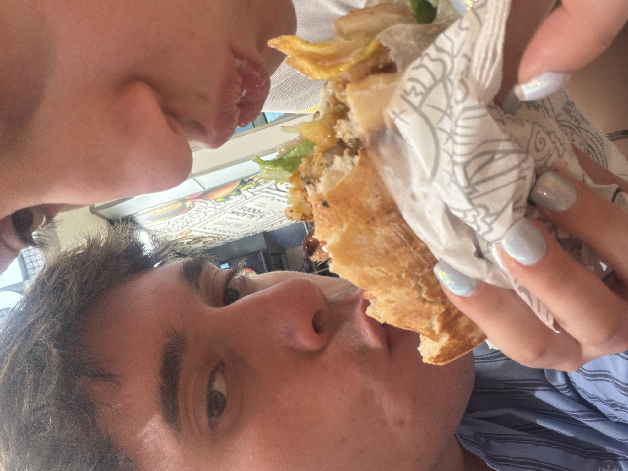 A photograph depicts two individuals sharing a sandwich, likely in Sofia, Bulgaria, though this cannot be definitively confirmed. The foreground features a person with brown hair and light skin, partially obscured by the sandwich. Their face is angled towards the camera, with their mouth open as if taking a bite. The sandwich itself is large, consisting of bread, lettuce, and other unidentified fillings, wrapped in patterned paper. It is being held by a hand with pale skin and light blue nail polish. Another person is positioned above, with only their lower face visible, also appearing to eat from the sandwich. The background includes a wall with writing on it, potentially advertising food items or menus. The scene suggests an intimate or casual sharing moment.