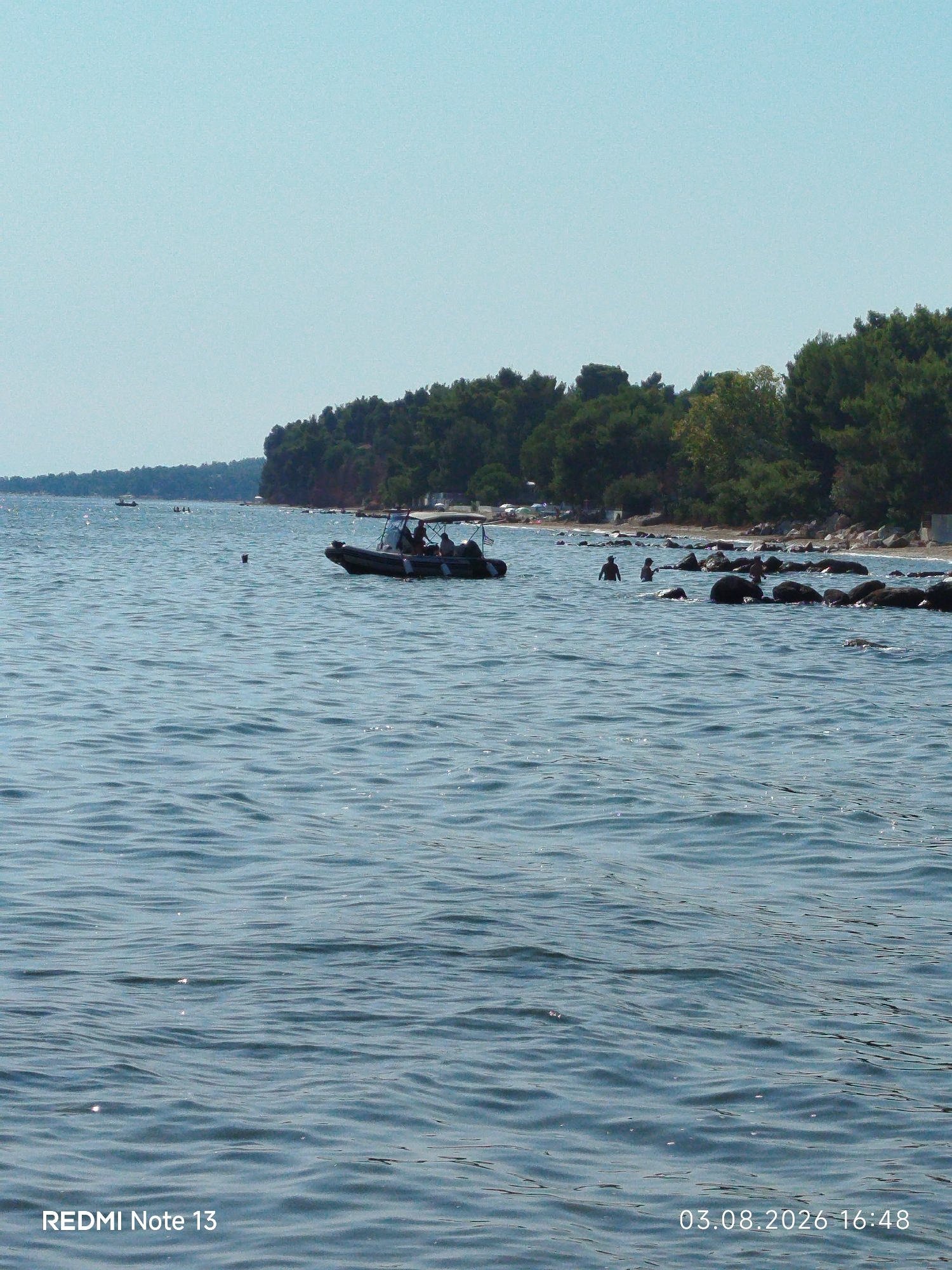 The image captures a serene coastal scene in Sithonia Municipal Unit, Greece, on August 3, 2026, at 16:48. The foreground is dominated by the calm, rippling blue water of the sea, with sunlight glinting off the surface. In the mid-ground, a small inflatable boat with a canopy is visible, carrying several individuals. Further in the distance, other small boats and people in the water can be observed. The background features a lush, tree-covered coastline, with a rocky shore interspersed with some vegetation. The sky is a clear, pale blue. The lower left corner of the image displays the text "REDMI Note 13," and the lower right corner shows the date and time.
