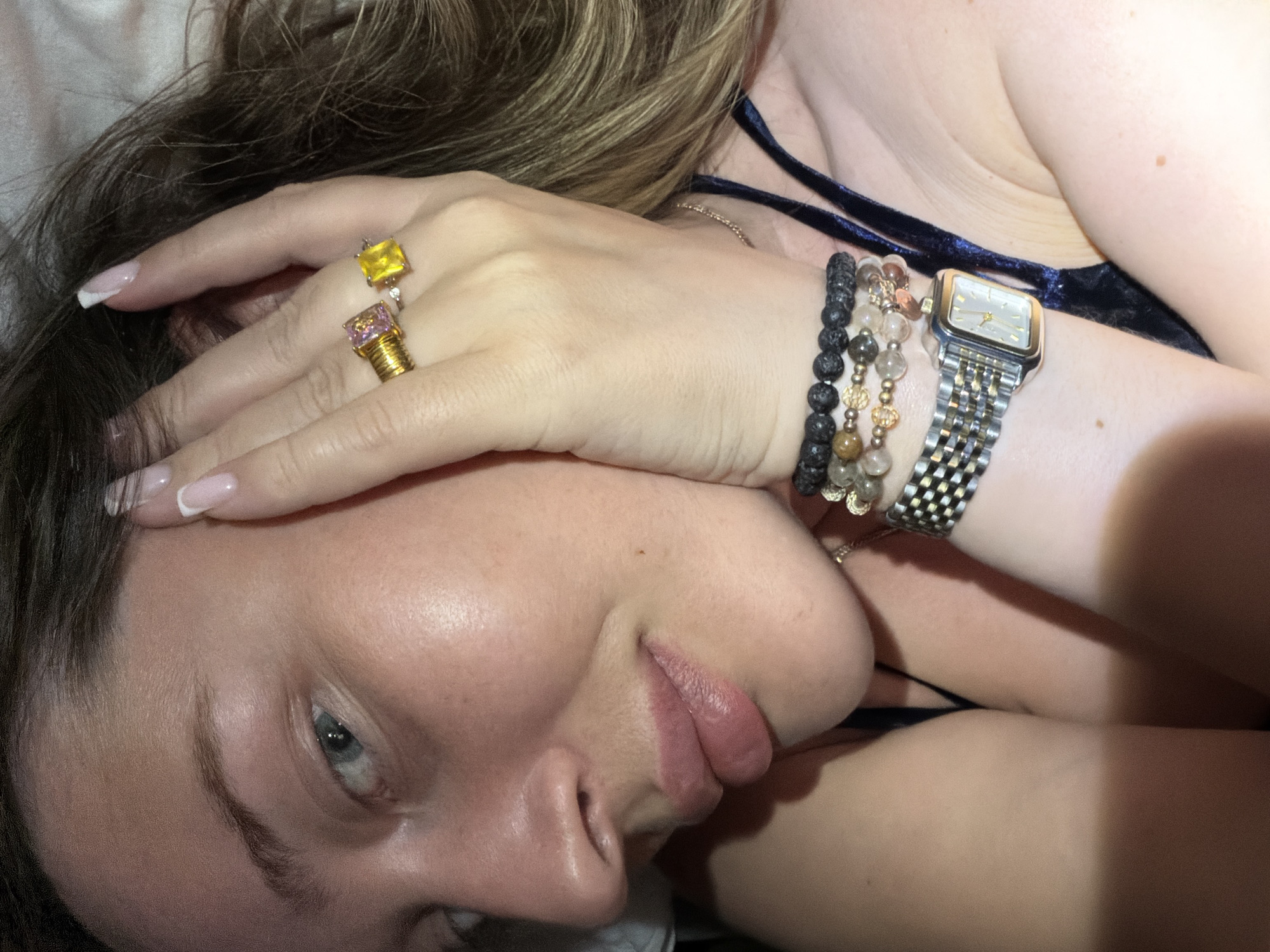 A woman lies on her back, head tilted to the left. She is wearing rings with square-cut yellow and pink stones on the third and fourth fingers of her right hand, which is partially covering her face. On her right wrist, she wears a metal watch and two beaded bracelets, one with dark, porous beads and another with lighter, translucent beads. She is wearing a dark, low-cut top, possibly velvet. The background is out of focus, suggesting a close-up shot. There is no information that indicates a specific location in Октябрьский, Russia.