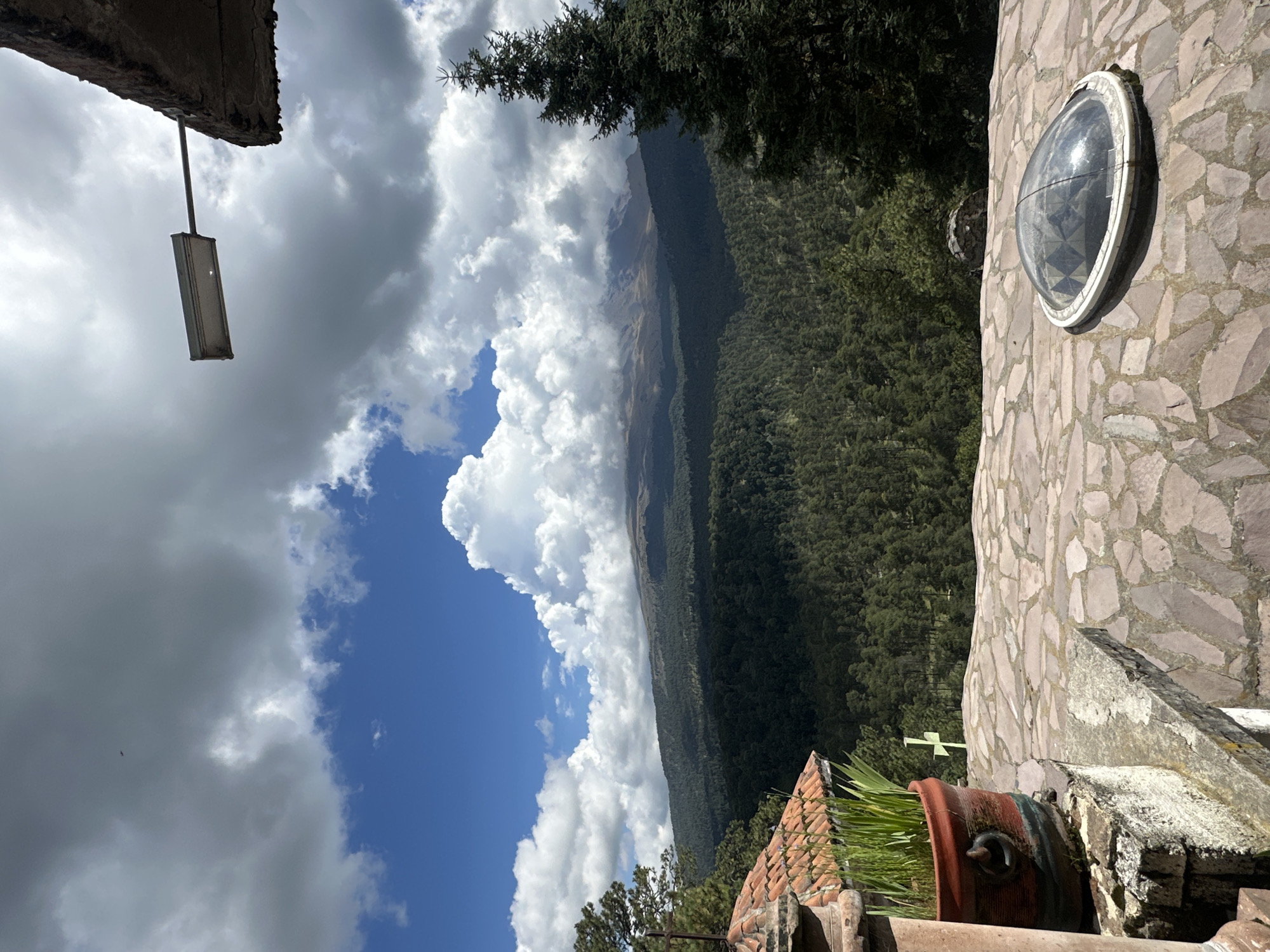 In San Mateo Ozolco, Mexico, a picturesque view unfolds featuring a verdant forest cascading down a mountainside towards a distant, mist-shrouded peak. In the foreground, a paved stone terrace offers a vantage point. To the left, a terracotta pot overflows with lush green grass, perched on a stone ledge. A cross is visible in the distance, amidst the trees. The sky above is a dramatic canvas of blue, dotted with large, billowing white clouds, some tinged with gray, suggesting a dynamic weather pattern. An outdoor light fixture is mounted on an overhang, facing the expansive landscape. The scene captures a serene, natural beauty, with architectural elements subtly integrated into the environment.