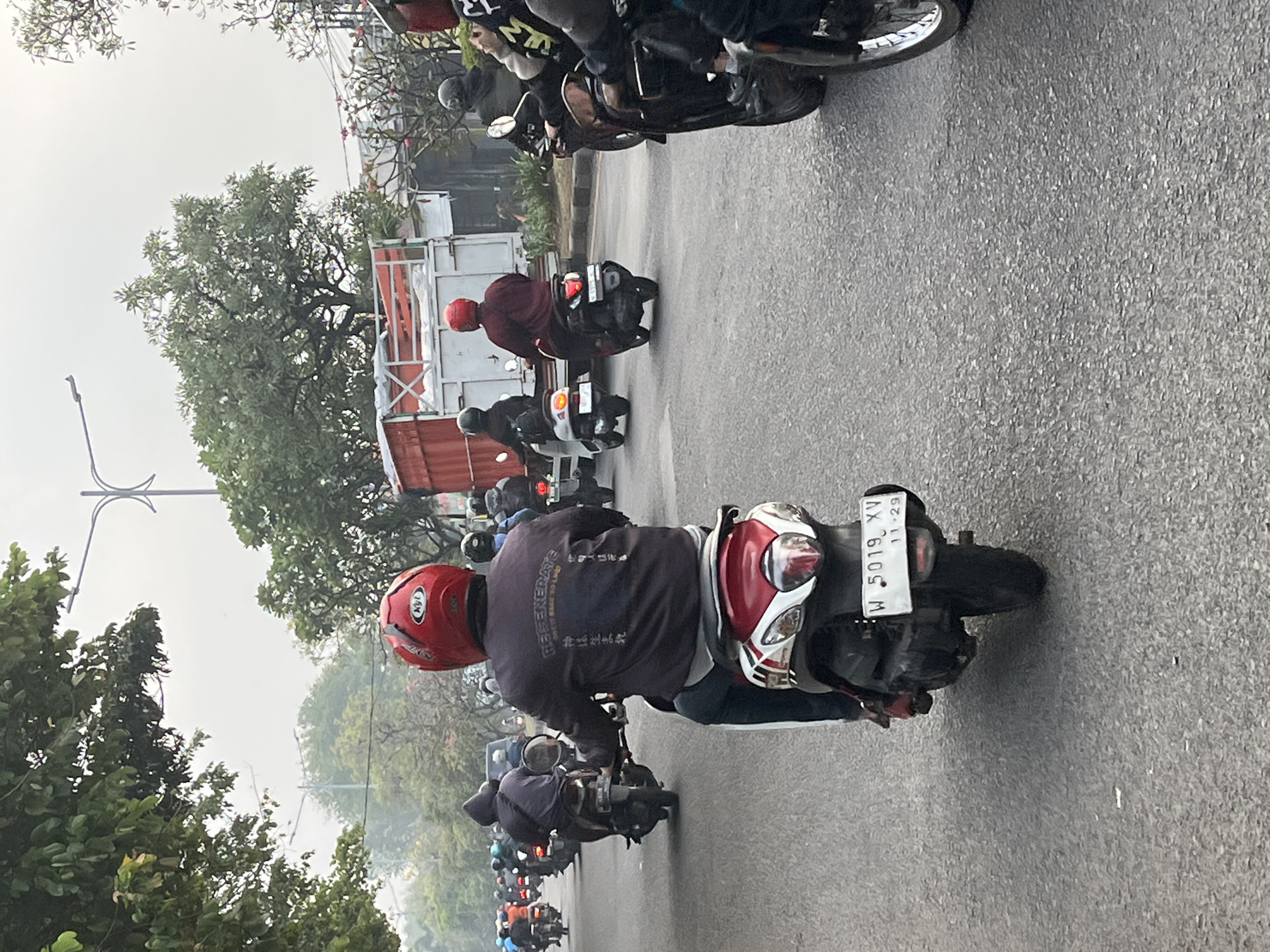 The image depicts a busy street scene in Sumorame, Indonesia, with a procession of motorcycles and scooters navigating the asphalt road. The foreground is dominated by a scooter with a red and white body, ridden by an individual wearing a red helmet and a dark jacket. The jacket has white text and Asian characters printed on the back. Behind this scooter, other motorcycles and riders are visible, some with helmets and casual attire, moving in the same direction.
In the mid-ground, a large red and white truck is parked or moving slowly on the right side of the road, partially obscuring other vehicles. Several more motorcycles are seen in front of the truck. The environment suggests a somewhat hazy or overcast day, with lush green trees lining the sides of the road and framing the sky in the background. Traffic lights and overhead wires are also visible, indicating an urban or semi-urban setting. The overall impression is one of daily commuting or transport in a populous area of Indonesia.