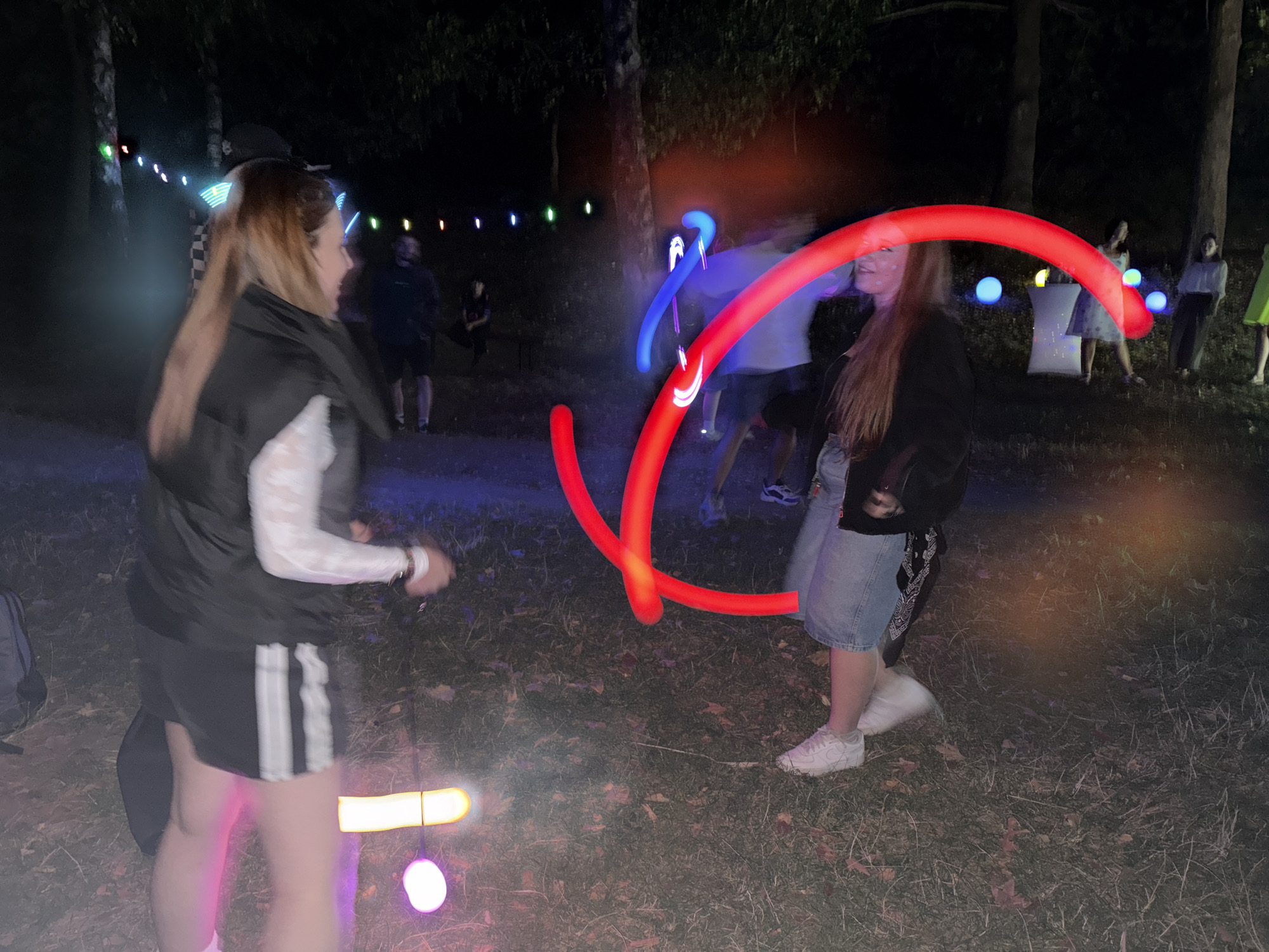 The image depicts an outdoor nighttime gathering with several people and illuminated objects. In the foreground, two individuals are prominently featured. The person on the left, with long blonde hair, wears a black vest over a white long-sleeved shirt and black shorts with white stripes. They are holding a light-up object. The person on the right, with long reddish-blonde hair, is wearing a black jacket and denim shorts. They are in the process of moving, with a large red light-up object forming a circular arc in front of them. Several other people are visible in the background, some standing and some sitting. The environment appears to be a wooded area with trees, and the ground is covered in dry leaves and grass. Various colored lights, including strings of small lights along trees and larger spherical lights on the ground, are scattered throughout the scene, contributing to a festive or event-like atmosphere. The image has a slightly blurred quality, likely due to the low light conditions and movement. The specific location in Zgorigrad, Bulgaria, is not visually identifiable from the provided image.
