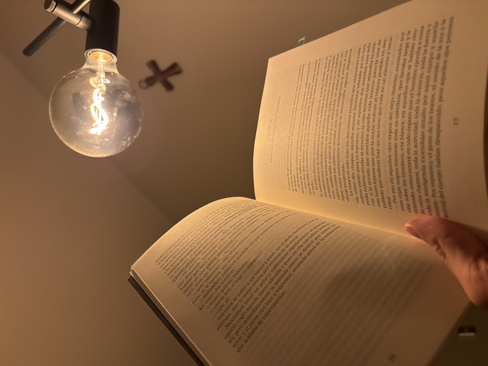 The image shows a book open, illuminated by an Edison-style light bulb hanging from above, with its filament visible. The bulb's warm glow casts shadows on the pages. The pages contain text in Spanish. A hand is holding the open pages. The background is out of focus, showing a decorative cross on the wall.