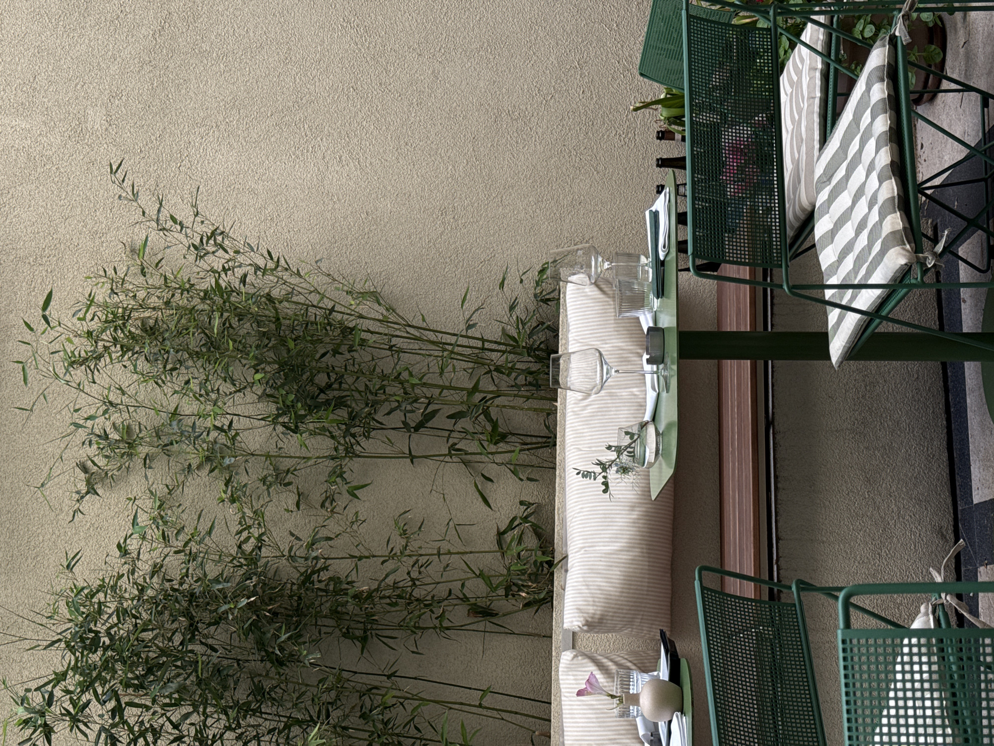 The image captures an outdoor dining scene, likely in Belgrade, Serbia, given the European architectural style. A table, draped with a striped cushion, is set with glassware, cutlery, and floral decorations. Green metal chairs, some with striped cushions, surround the table. Lush bamboo foliage lines the background, obscuring a textured wall. Several wine bottles stand near the table. The scene suggests a relaxed, possibly upscale, dining environment.