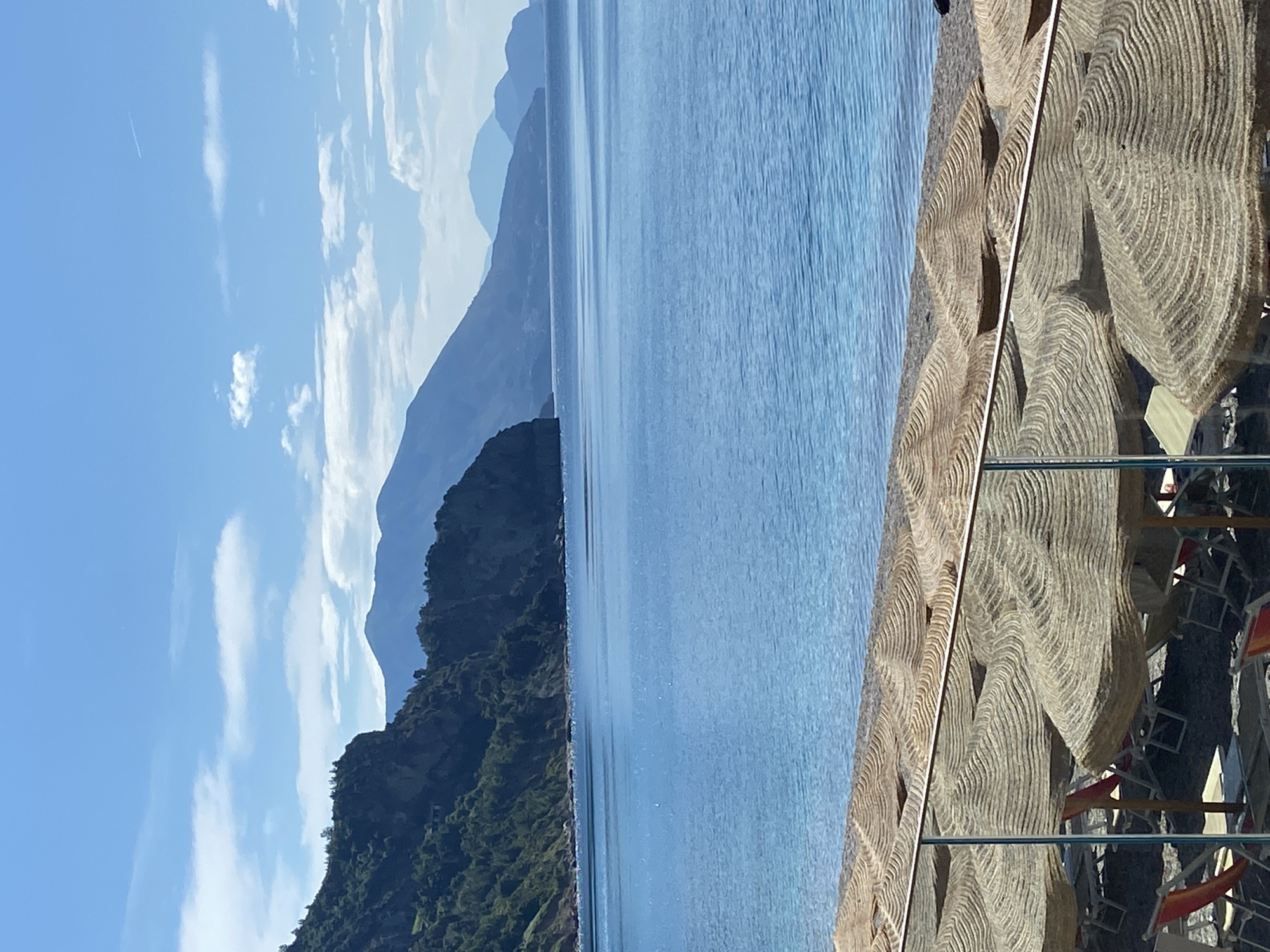 The image depicts a tranquil scene in Prijevor, Montenegro. A body of water stretches between a forested, rocky coastline to the left and beach umbrellas lining a shore to the right. Distant, hazy mountains fade into the background. The sky above is blue with scattered clouds. The beach umbrellas are made of natural material. Reflections of beach chairs can be seen in the foreground glass.