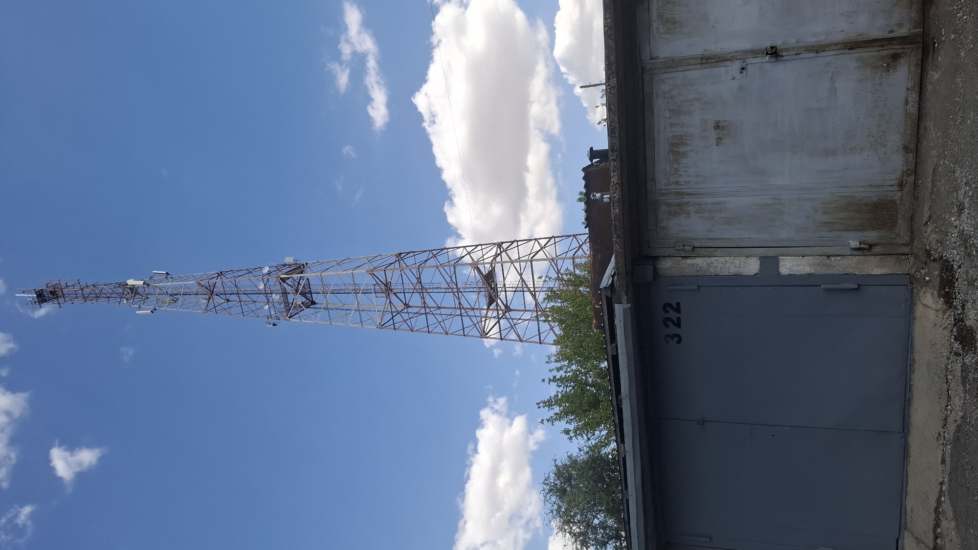 A cellular tower stands tall against a bright blue sky with scattered white clouds. The tower is a lattice-like metal structure, with several antennas and dishes mounted at various levels. In the foreground, there is a row of garages. The left garage door is painted dark gray and has the number "322" stenciled in black. The adjacent garage door to the right is older, with a distressed metallic finish and visible rust and grime. Trees are visible behind the garages, partially obscuring the lower portion of the cellular tower. The ground in front of the garages is a mix of asphalt and gravel. This scene is located in the city of Shymkent, Kazakhstan.