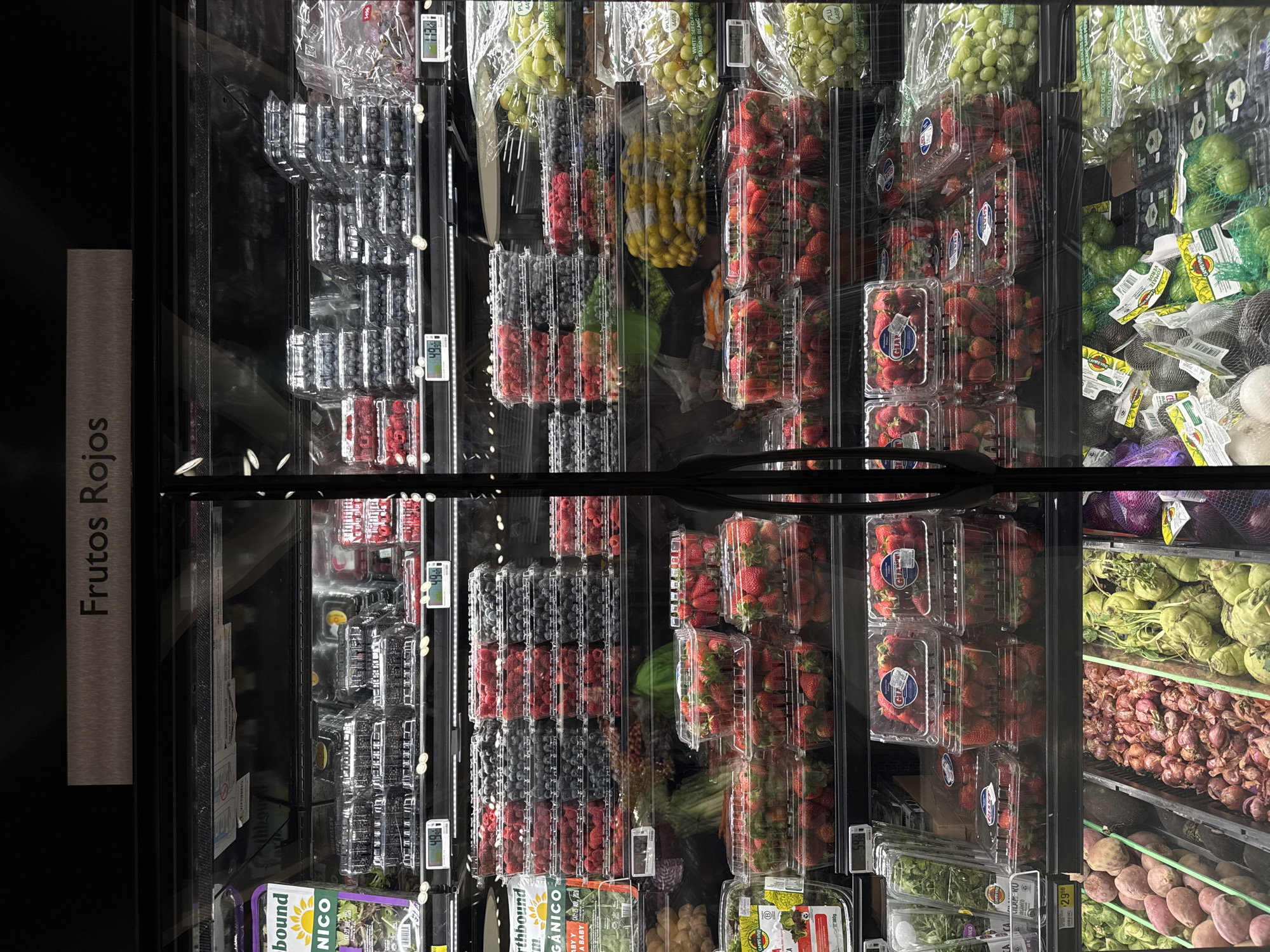 The image shows a refrigerated display case at a grocery store in Puebla, Mexico. The sign on the left indicates "Frutos Rojos" (Red Fruits), which is supported by the produce inside the case. Packaged strawberries and assorted packaged berries dominate the middle shelves. Further to the left, there are blueberries and raspberries in clear plastic clamshells. To the right of the strawberries, there are green fruits and some bulb vegetables. Digital price tags are visible along the vertical edges of the shelves. The scene appears to be inside a grocery store.