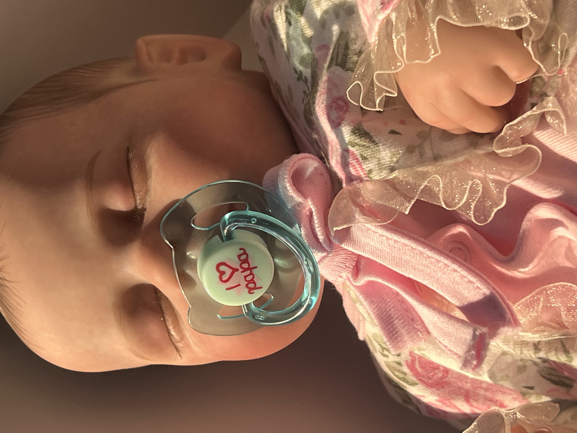The image shows a close-up of a baby doll with a pacifier in its mouth. The doll is wearing a pink outfit with a floral pattern. The background is a dark brown color. There is no indication of location in Votkinsk, Russia.