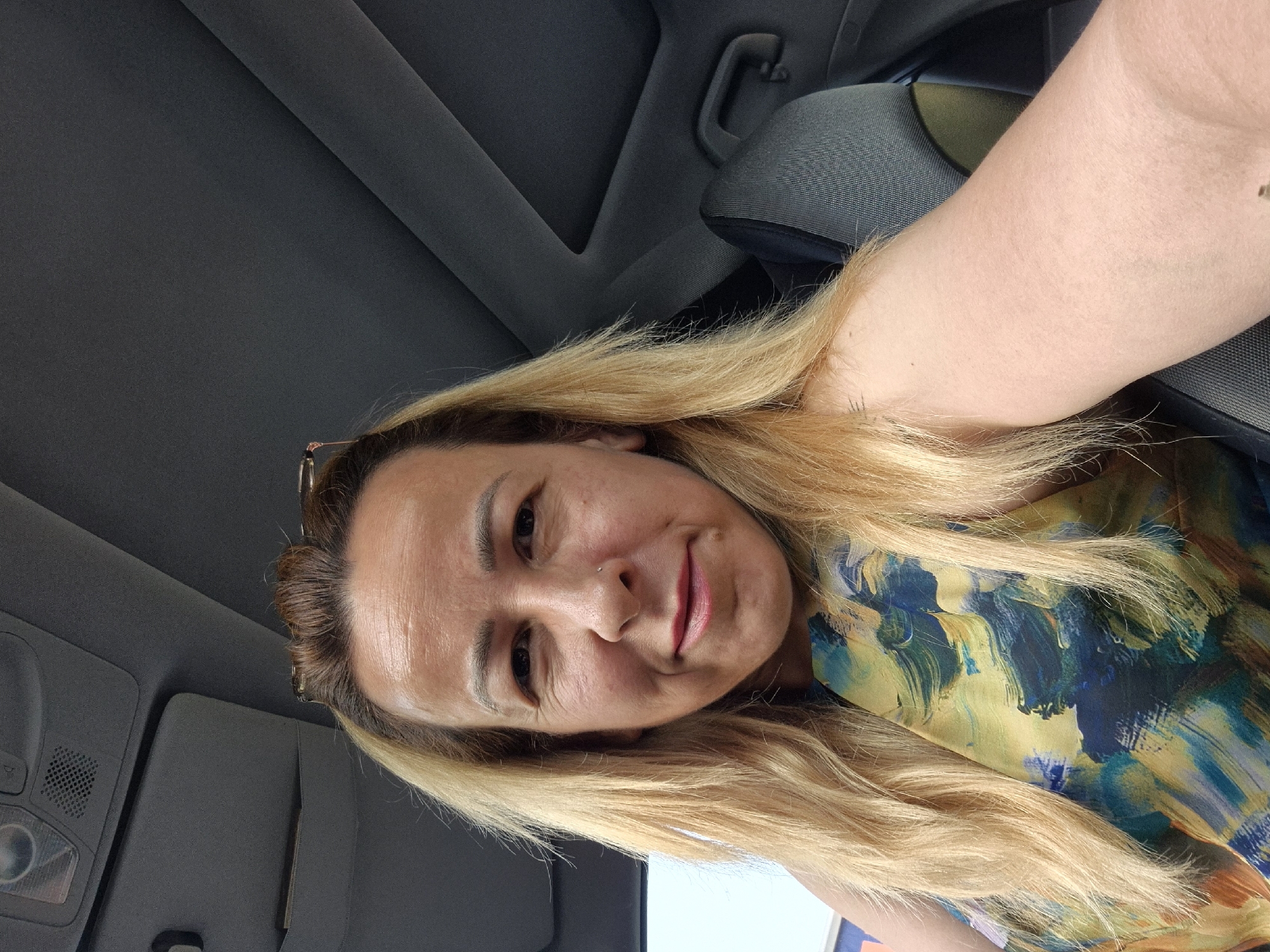 A woman with blonde hair and a floral-patterned top is sitting in a car. She is looking at the camera and smiling. The car's interior is visible in the background, including the ceiling and seats.