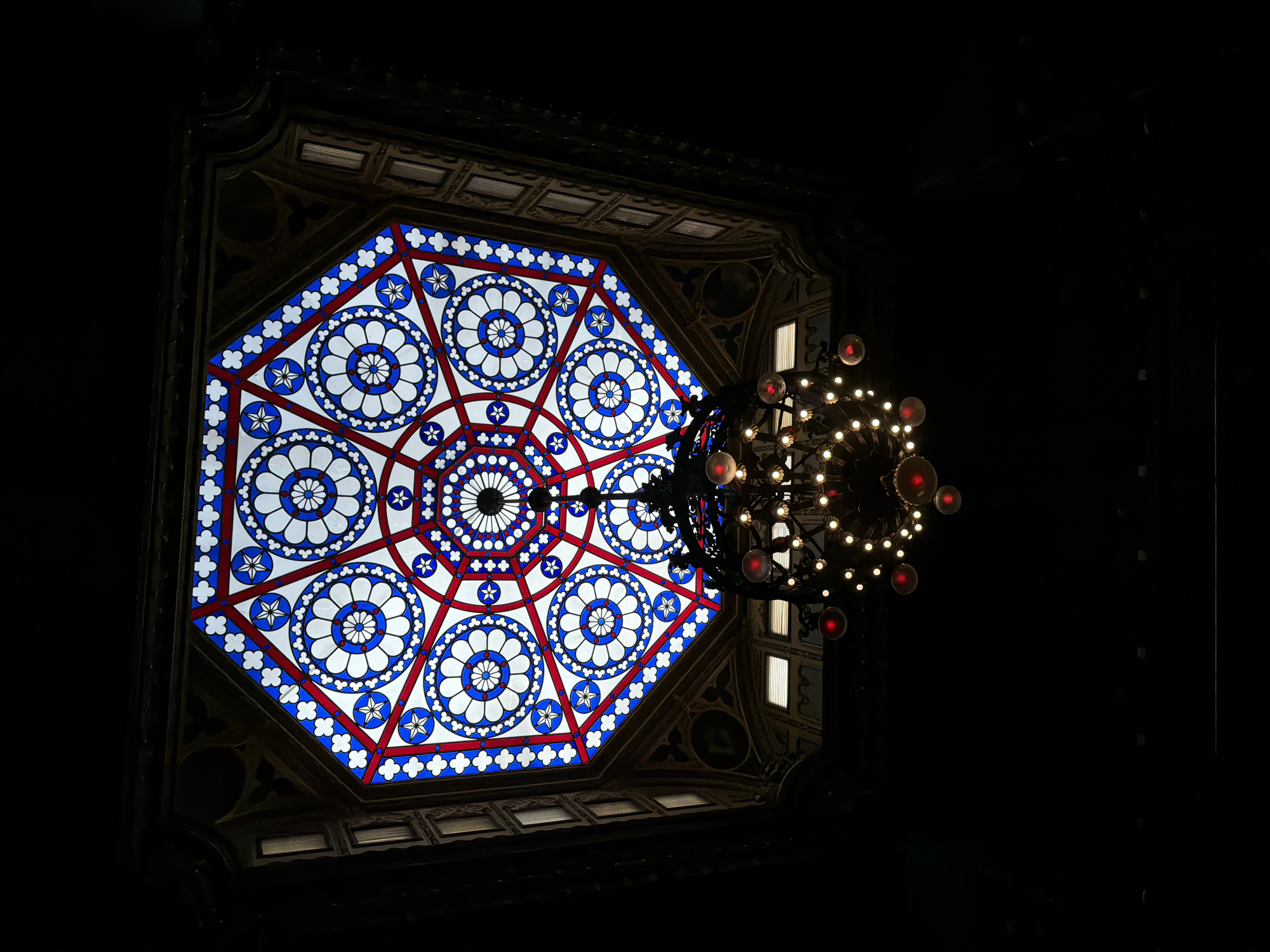 The image shows a stained-glass ceiling with an octagonal design featuring intricate geometric patterns in red, white, and blue. A dark, ornate chandelier hangs from the center of the ceiling, illuminated by numerous lights, some of which have red-tinted bulbs. The ceiling is framed by dark architectural elements, suggesting an indoor space. The lighting is dim, with the stained-glass and chandelier being the primary sources of illumination. There are no people visible in the image.
This scene is located in the **Museu Histórico Nacional (National Historical Museum)** in Rio de Janeiro, Brazil. The stained-glass ceiling is a prominent feature within the museum's architecture.