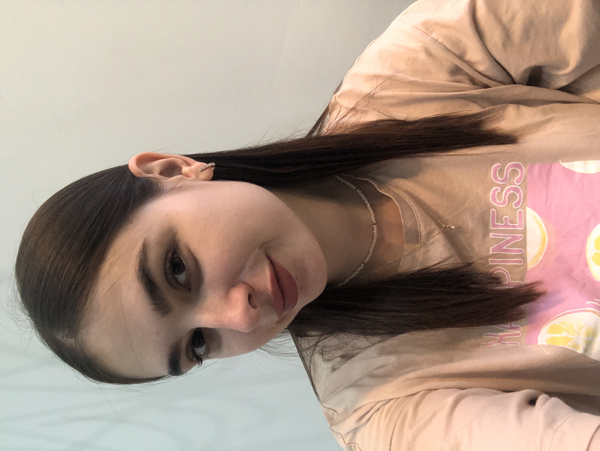 A young woman with long, dark brown hair and fair skin is photographed from the chest up in a selfie. She is wearing a light tan t-shirt with pink lettering that reads "HAPPINESS" and a graphic of sliced lemons. She also wears a delicate pearl necklace and a small, sparkling earring in her left ear. Her skin is clear, and she has subtle makeup on, with lipstick in a muted berry shade. The background is a plain, light-colored wall, suggesting an indoor setting, possibly a residence in the city of Chaikovsky, Russia. There are no other people or objects of significance in the frame. The lighting is soft and even, illuminating her face and upper body.