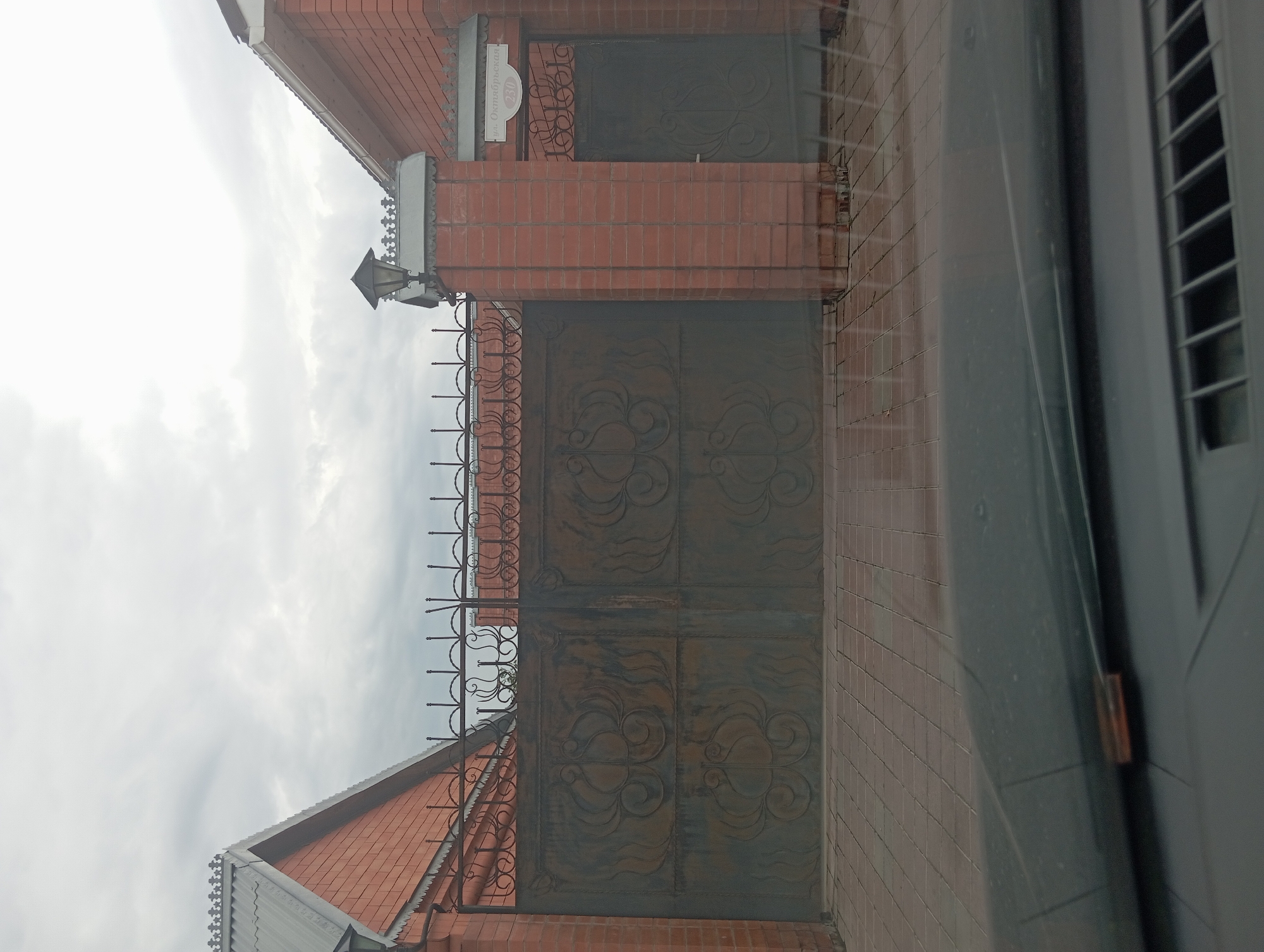 The image shows a brick house with a metal gate. The gate is made of dark metal with ornate designs. There is a light fixture on top of a brick pillar next to the gate. A sign with Russian writing is visible on the pillar. The ground in front of the gate is paved with bricks. The sky is overcast. The image appears to be taken from inside a vehicle, as the dashboard and windshield are visible in the foreground.