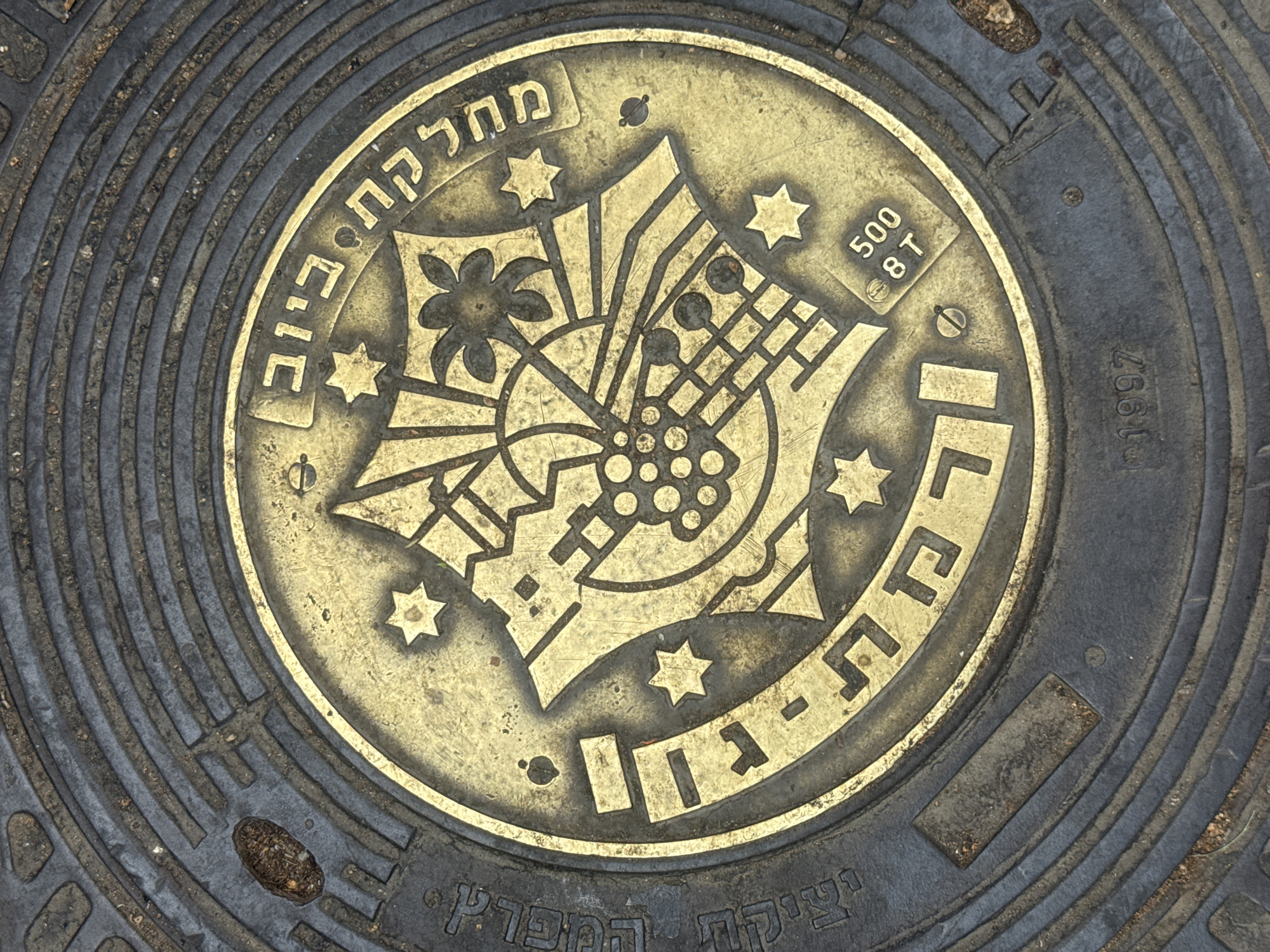 The image shows a close-up of a manhole cover, likely located in the city of Ramat Gan, Israel, as inferred from the Hebrew text inscribed on it. The cover is circular, made of metal, and appears aged and weathered. The central design consists of a stylized emblem featuring a floral motif, geometric shapes, and several six-pointed stars, all rendered in a golden or bronze-colored metal against a darker background. Hebrew text is arranged in an arc along the upper and lower rims of the central design. The numbers "500" and "8T" are visible near the upper right. The number "1997" is also visible, likely indicating the year of manufacture. The surrounding frame of the manhole cover exhibits concentric rings, with evenly spaced rectangular cutouts.