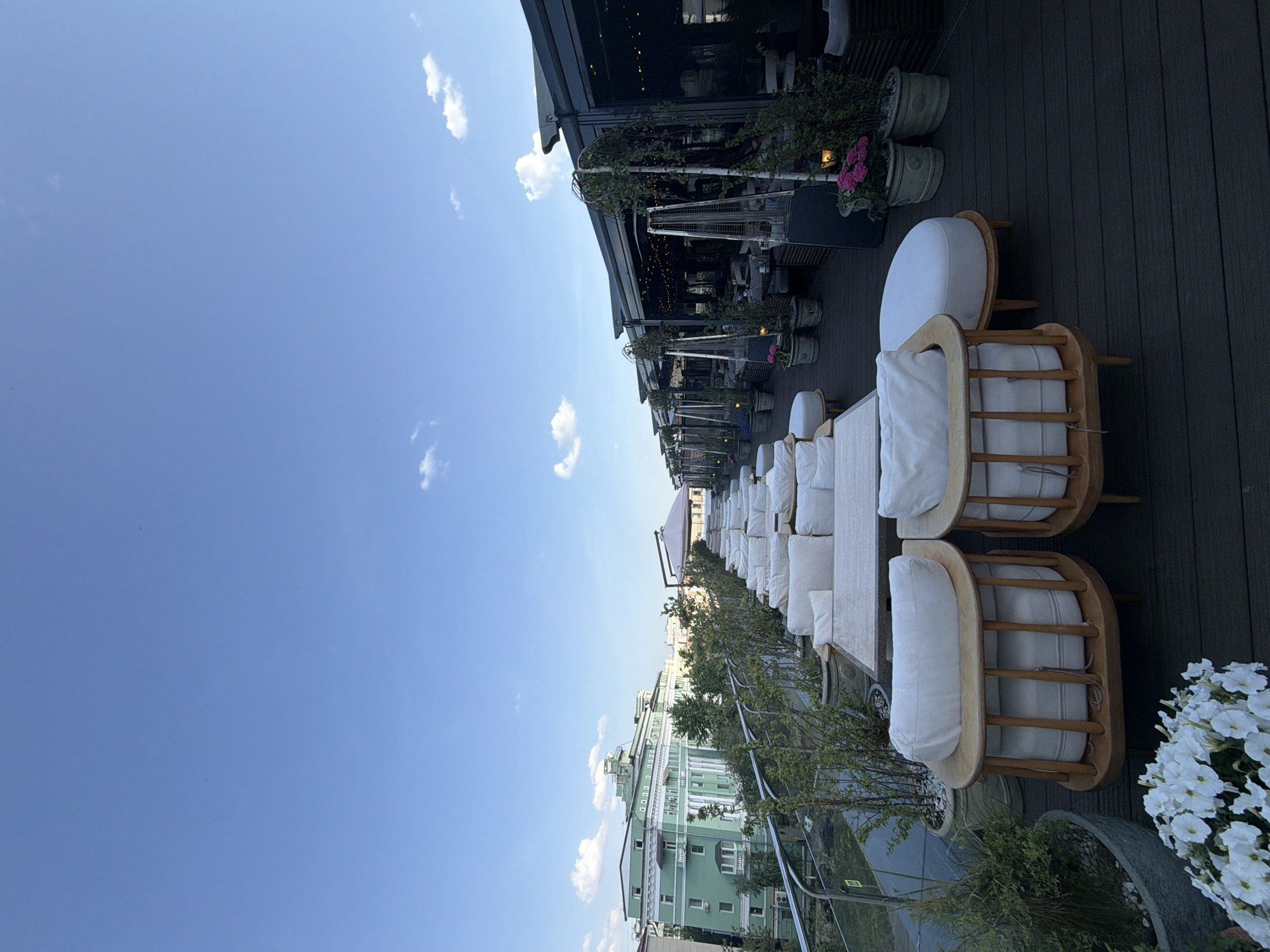 The image depicts a rooftop terrace, likely in Kazan, Russia, with several lounge chairs and sofas upholstered in white cushions arranged in a row. The terrace has a wooden deck and a railing along the edge. Potted plants, including trees and flowers, are placed between the seating areas and along the railing. A building with a light green facade can be seen across the street, indicating an urban setting. The sky is blue with scattered clouds. Part of a building structure with awnings and what appears to be a restaurant area is visible at the top of the image.