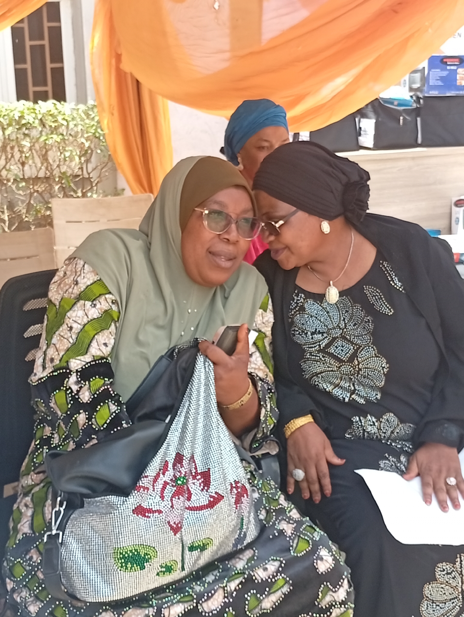 Two women are seated and engaged in conversation at what appears to be an outdoor or semi-outdoor social event in Unguwar Sarki, Nigeria, bathed in bright daylight. The woman on the left, wearing a light olive green hijab and a dress with patterned sleeves in green, white, black, and gold, holds a mobile phone in her right hand, seemingly showing something to her companion. She wears glasses and a gold bracelet, and a distinctive silver sequined handbag with a red and green floral design rests on her lap.
The woman on the right, clad in a black hijab and a black dress adorned with intricate silver beaded patterns, leans in attentively, looking towards the phone. She wears glasses, a gold pendant necklace, gold earrings, a gold bracelet, and multiple rings. Both women appear relaxed and engaged.
In the background, a third woman wearing a blue head covering is partially visible between the two main subjects. The setting includes orange draped fabric overhead, lush green foliage behind the woman on the left, and a building with a window. To the right, various packaged goods or appliances are visible on shelves or a table, suggesting a display or market setup. There is no discernible text visible in the image.