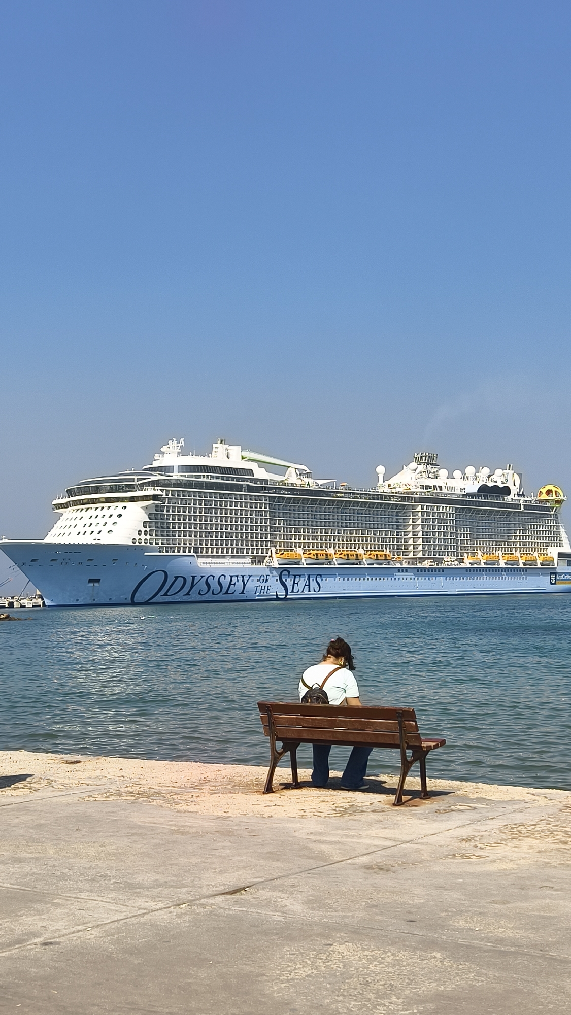 An outdoor, vertical, medium shot captures a person sitting on a wooden bench facing a large cruise ship docked in a body of water. The person, viewed from behind, has dark hair and wears a light-colored t-shirt and dark pants, with a black backpack on. The cruise ship, "Odyssey of the Seas," is prominently displayed, dominating the background. The ship is white with blue lettering and yellow life rafts visible along its side. The sky is clear and blue, and the water is calm. The foreground consists of a concrete walkway leading to a sandy area where the bench is situated. The image was taken in Kuşadası, Turkey, a popular port city known for its cruise ship terminals.