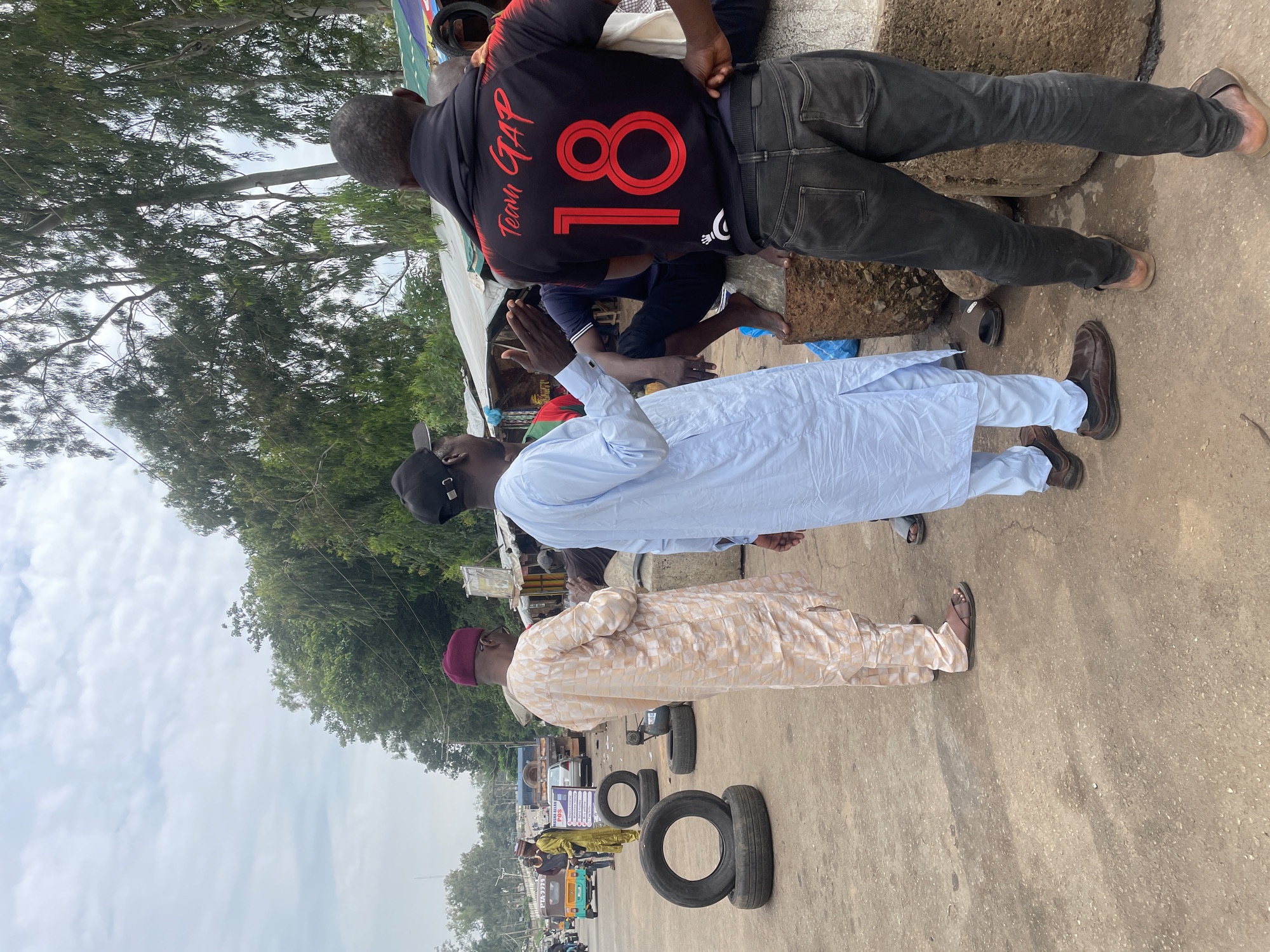 The image depicts an outdoor scene in Jos, Nigeria, likely a marketplace or roadside area, characterized by a paved surface and scattered objects. In the foreground, four men are visible. One man, wearing a light blue traditional outfit and a black cap, is facing away from the camera and gesturing with his right hand, appearing to be in conversation. To his left, another man in a light-colored, patterned traditional garment and a maroon cap is also facing away, looking down. To the right of the man in blue, a third man, wearing a black shirt with "Team GAP 18" printed on the back and dark denim jeans, is seen from behind. He is standing near a large, rough-hewn stone. A fourth man, partially obscured, is visible to the far left of the frame, wearing a yellow traditional outfit and a hat, standing near a sign that reads "MADAM ALLAN POS".
Several discarded tires are scattered on the ground, some stacked on top of each other. In the background, various stalls with awnings are set up, suggesting commercial activity. Trees with dense foliage line the upper portion of the image, and a cloudy sky is visible above. Vehicles, including a tricycle-like vehicle, and a truck, are present on a road in the distance. The overall impression is a busy, perhaps informal, commercial hub.